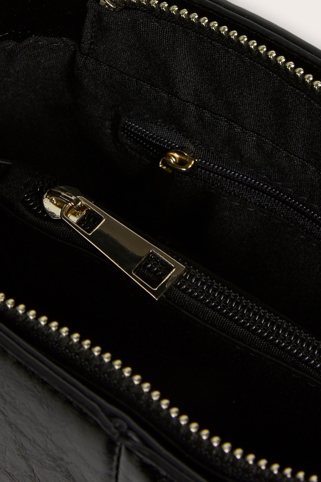 Lähikuva mustan tekonahkaisen käsilaukun sisältä, jossa on kullanväriset vetoketjut ja musta vuori.