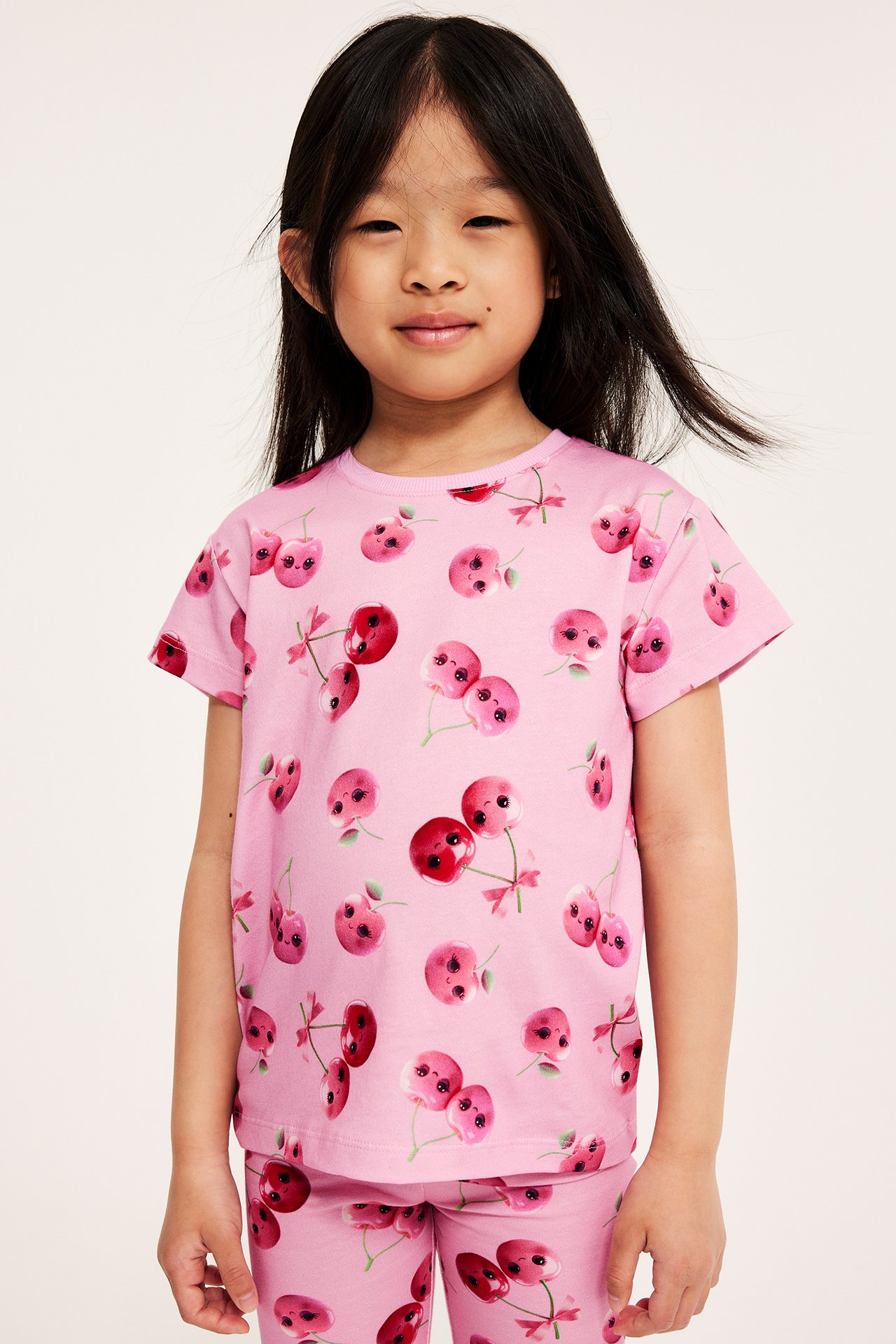 Framifrån, ett barn i rosa bomulls-t-shirt med körsbärstryck och matchande rosa byxor.