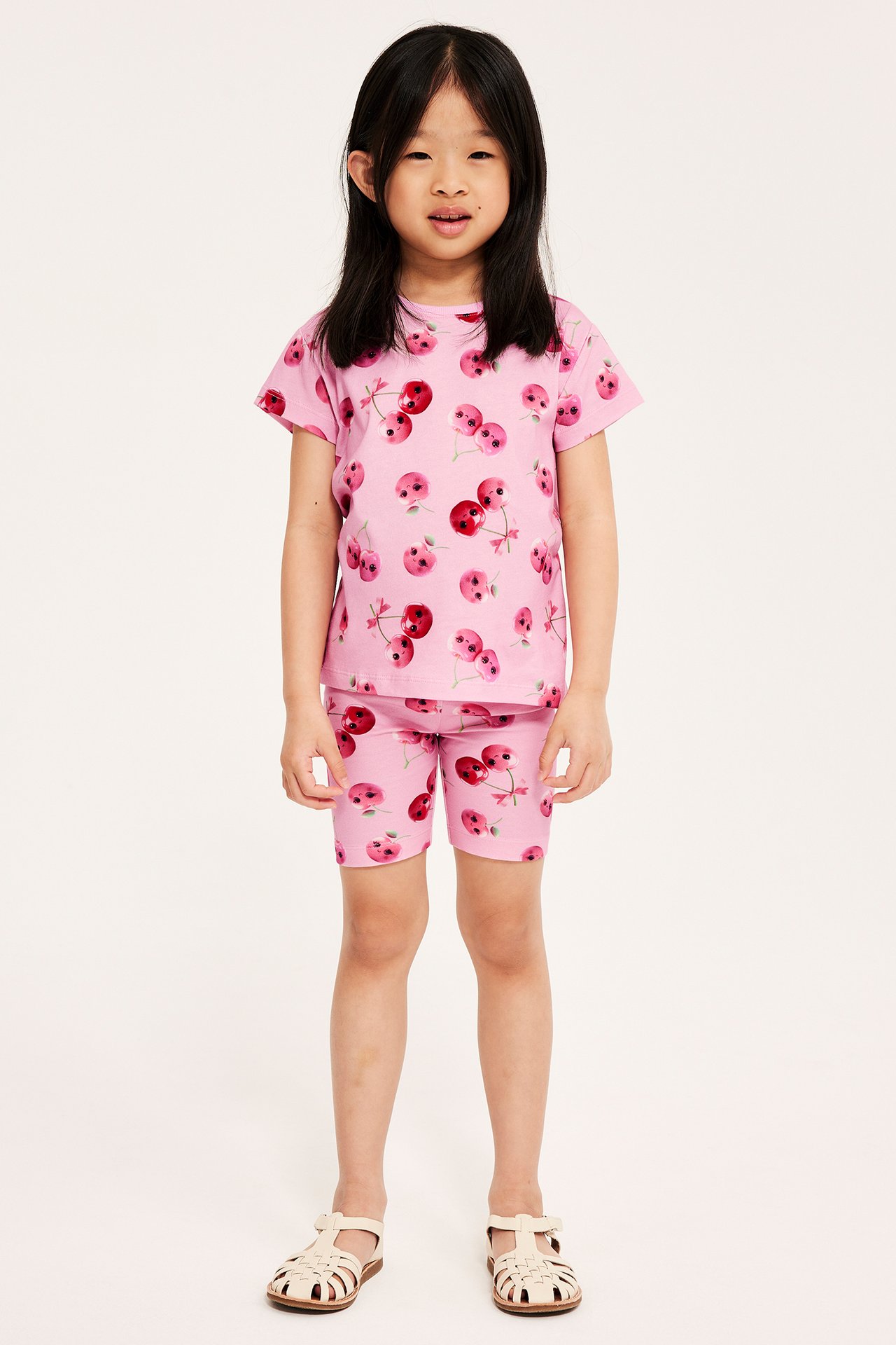 Framsida av ett barn i rosa bomulls-t-shirt med körsbärstryck, matchande shorts och ljusbeige sandaler.