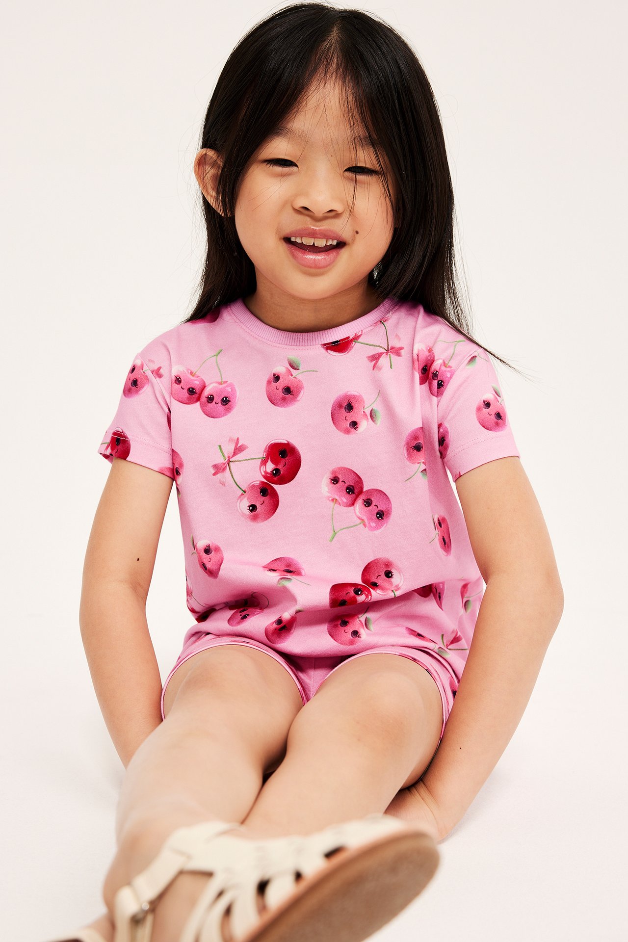 Framifrån, ett barn i en rosa bomulls-t-shirt med körsbärstryck och matchande shorts.