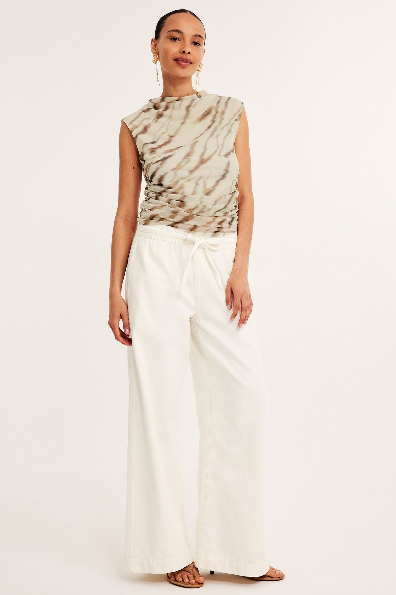 Frontbilde av kvinne i mønstret beige mesh-topp med rynker, hvite vide bukser og brune sandaler.