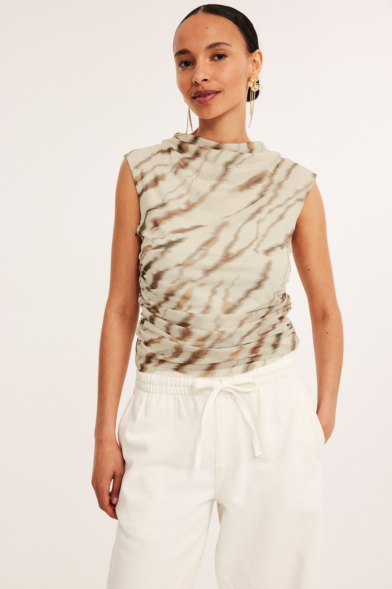 Forfra: kvinne i beige mønstret meshtopp med drapert hals og hvite vide bukser.