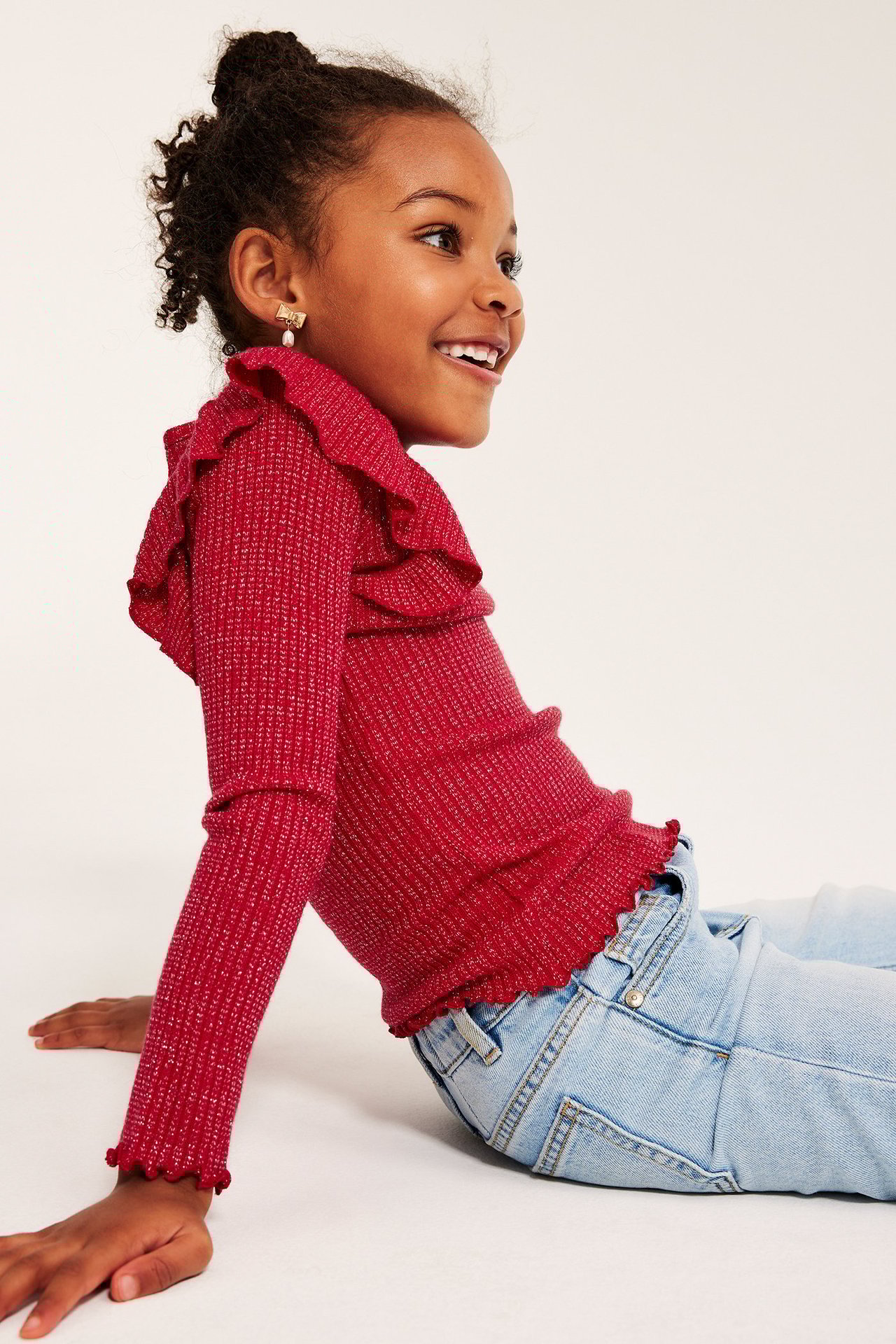 Flicka i röd, glittrig, ribbad topp med volanger, långärm, sittande på golvet i jeans.