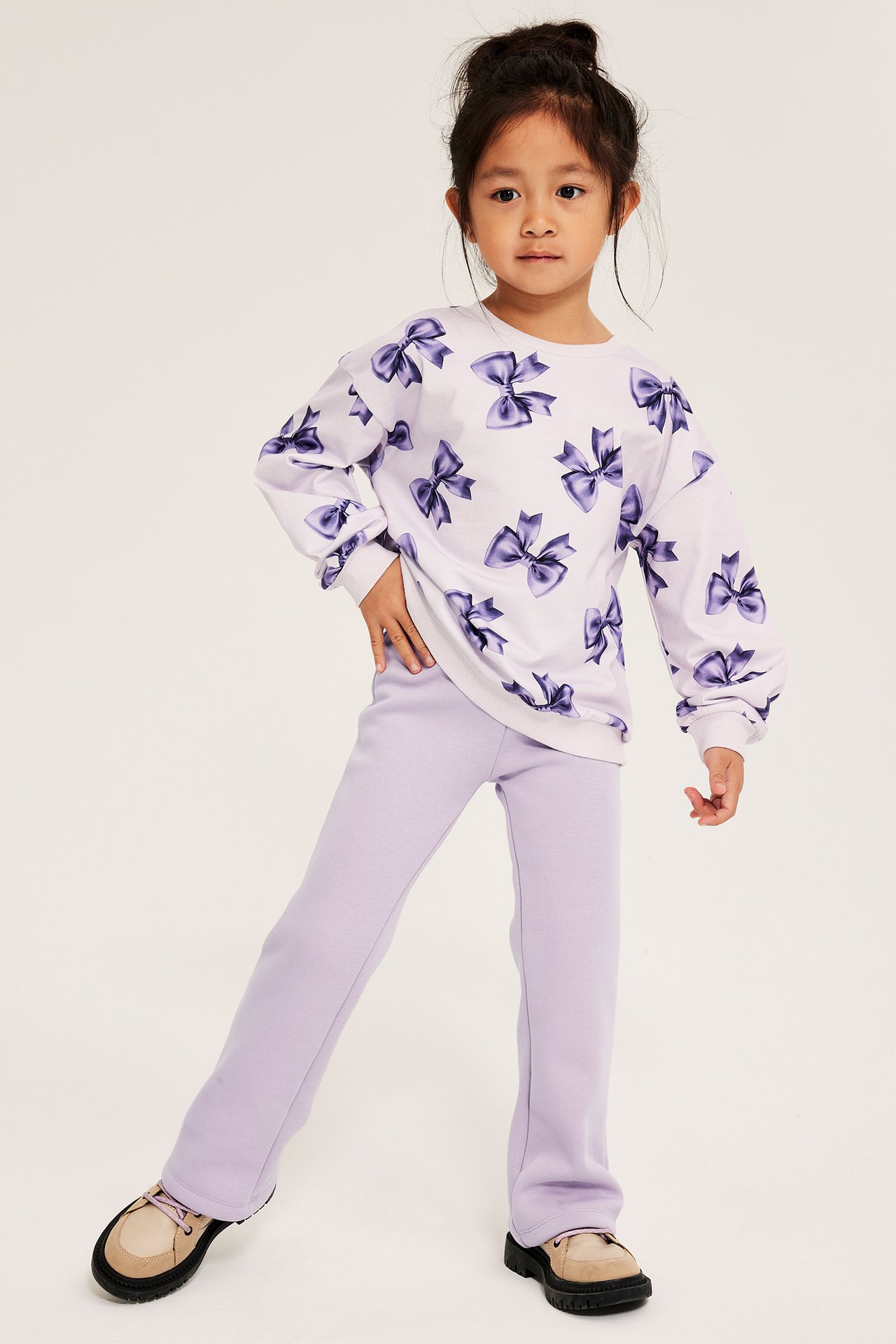 Nuori tyttö valkoisessa hupparissa tumman lilalla rusetti-kuviolla ja vaalean lilalla flare-leggingsillä.