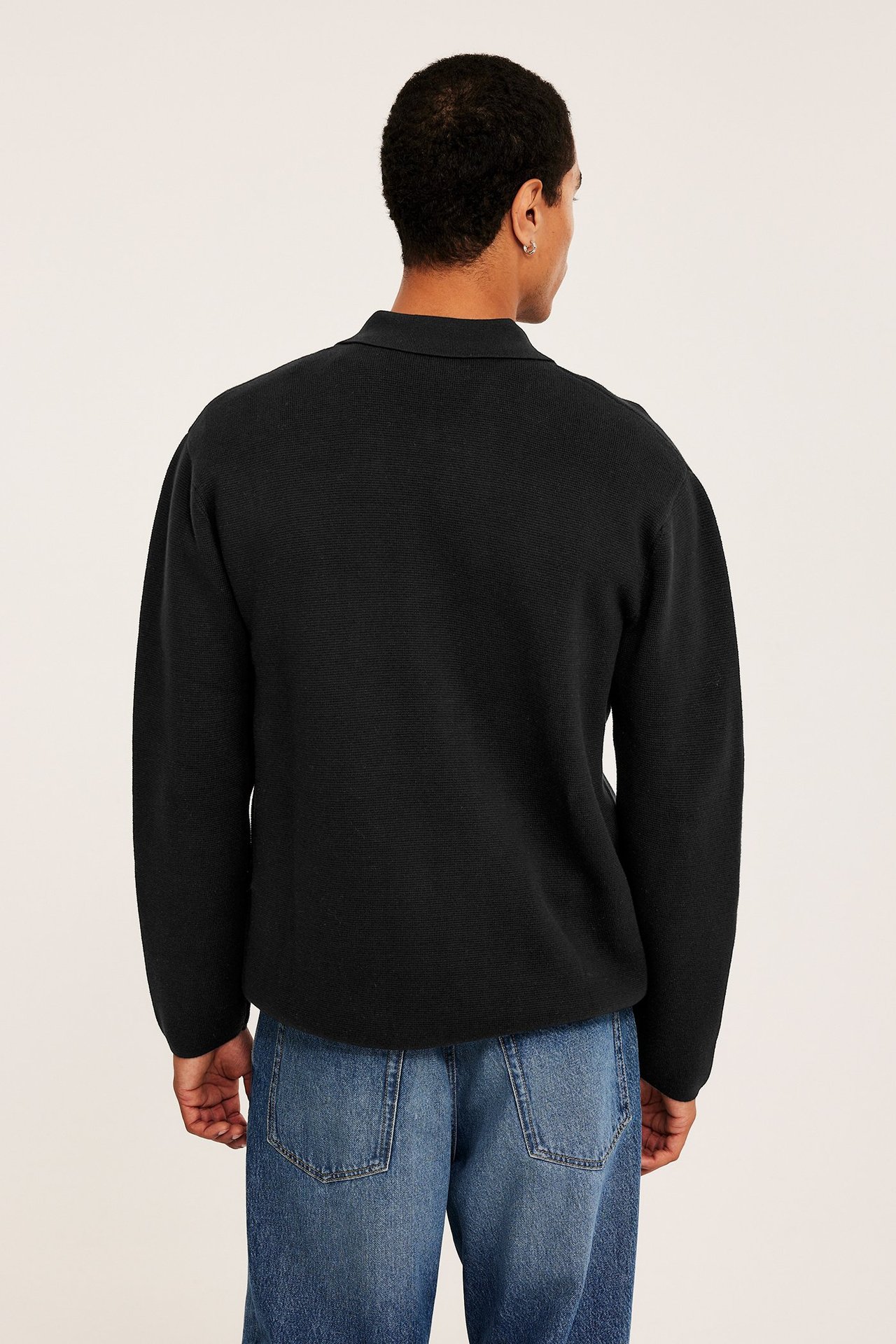 Ryggvy av en man i svart stickad bomullströja med krage, kombinerad med blå jeans.