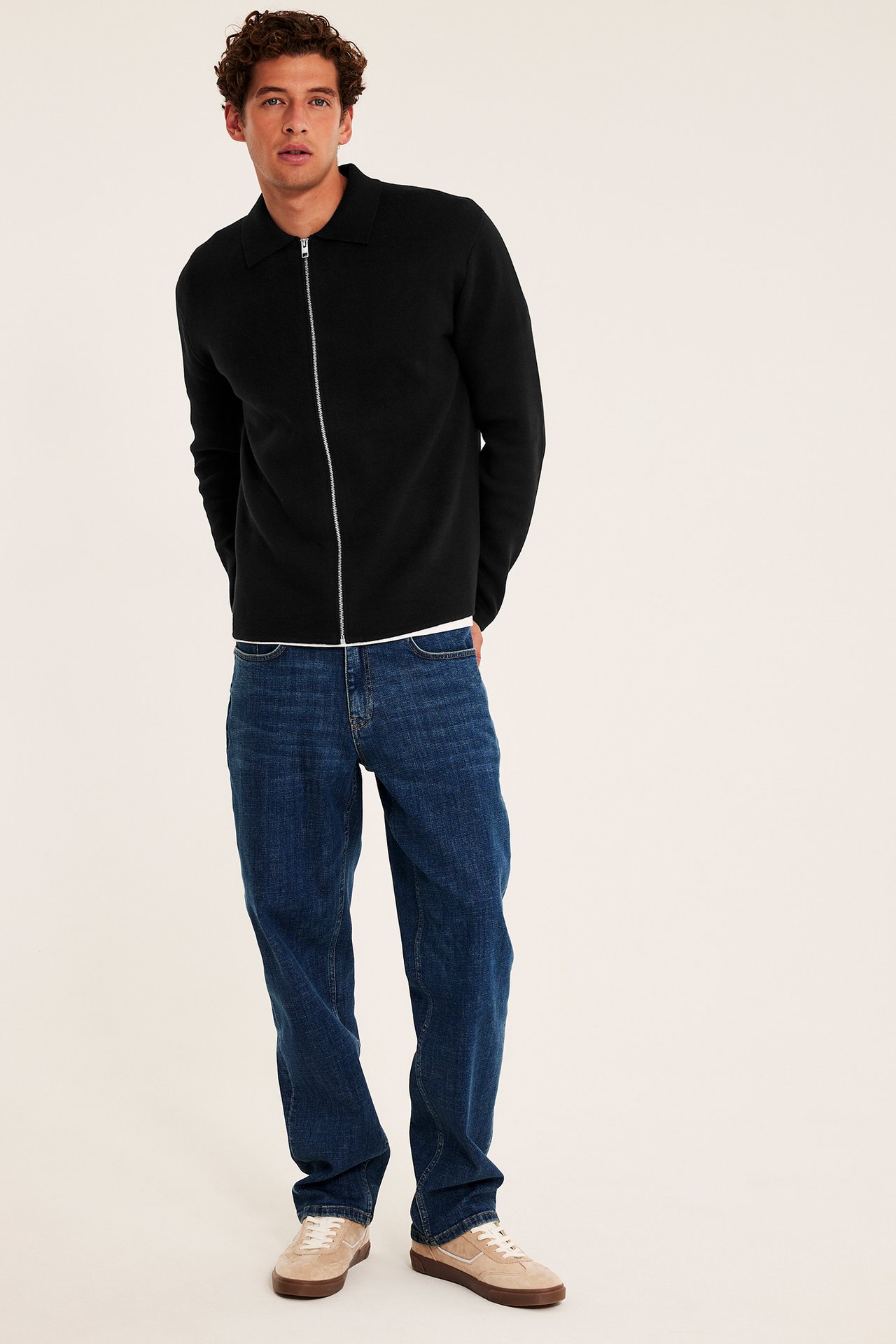 Frontbilde av en mann i svart kragejakke med glidelås, blå jeans og beige joggesko.