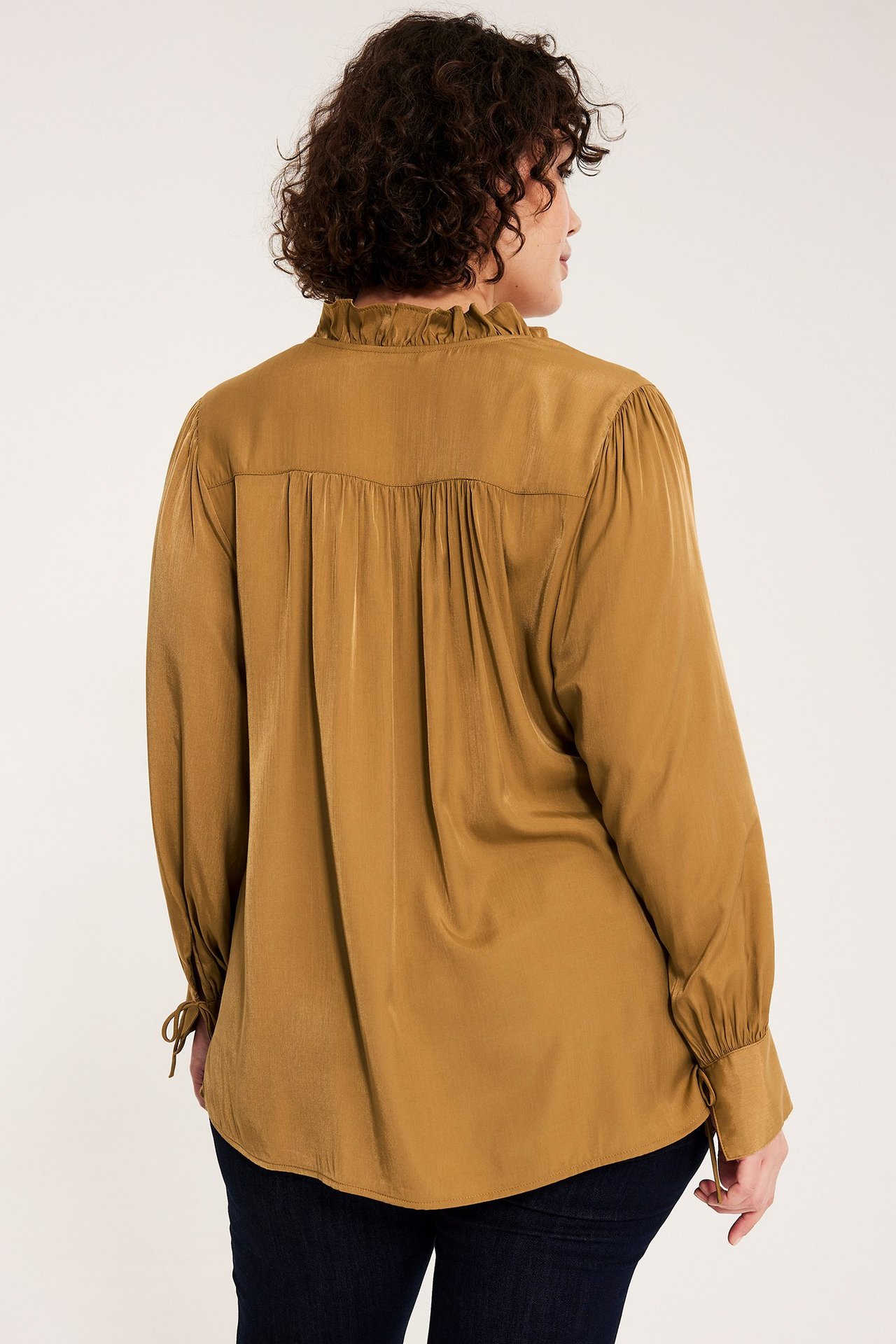 Naisen takakuva yllään beigessä pitkähihaisessa puserossa, jossa röyhelökaulus ja sidottavat hihansuut.