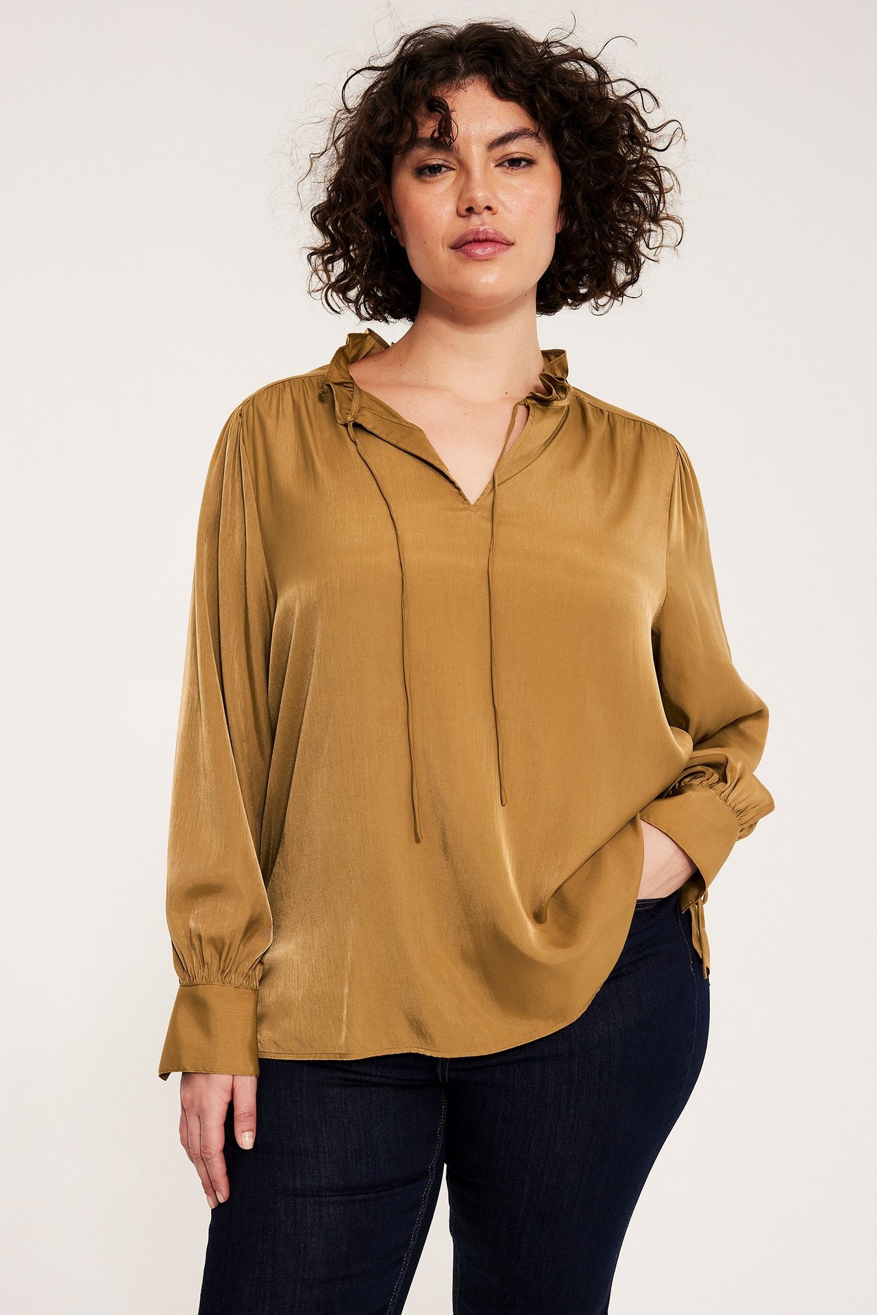 Framsida av kvinna i lös beige långärmad blus med volang och knytdetaljer, samt mörkblå jeans.