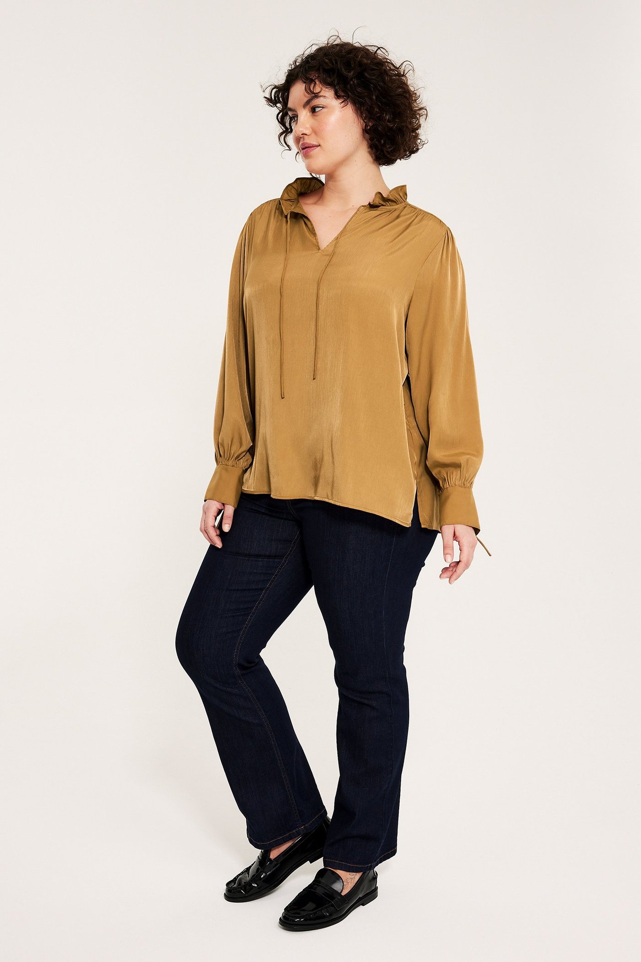 Sivukuva: nainen beigessä pitkähihaisessa röyhelökauluspuserossa, tummissa farkuissa ja mustissa loafer-kengissä.