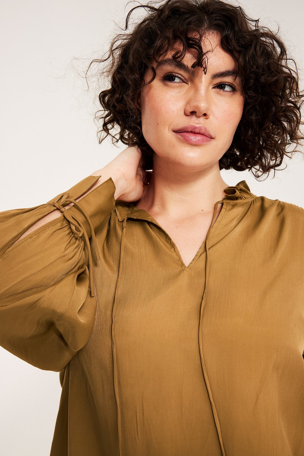 Lähikuva naisesta beigessä pitkähihaisessa puserossa, jossa röyhelökaulus ja solmittavat yksityiskohdat.