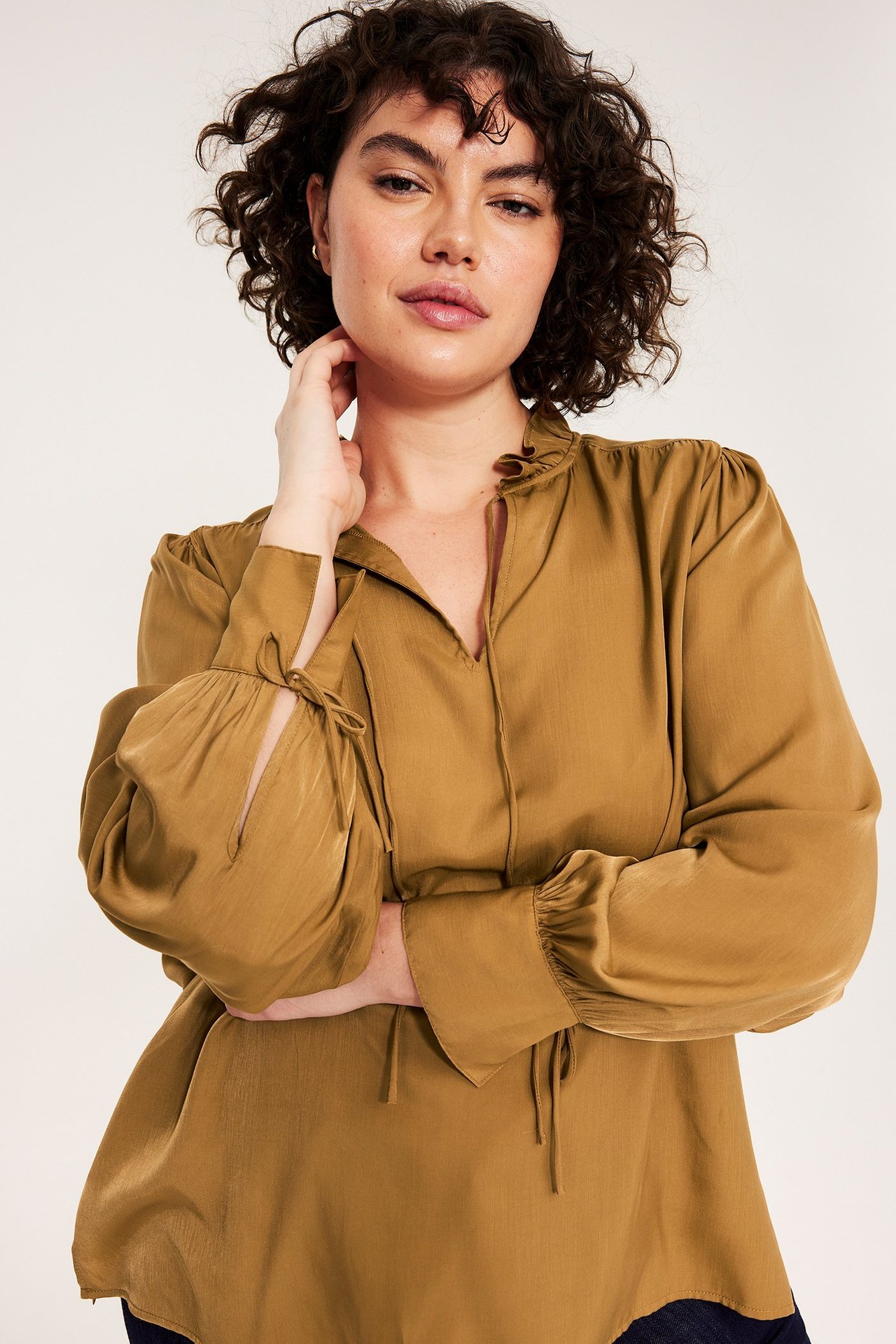 Widok z przodu kobiety w beżowej bluzce z długim rękawem, falbanką przy dekolcie i wiązaniami przy mankietach.