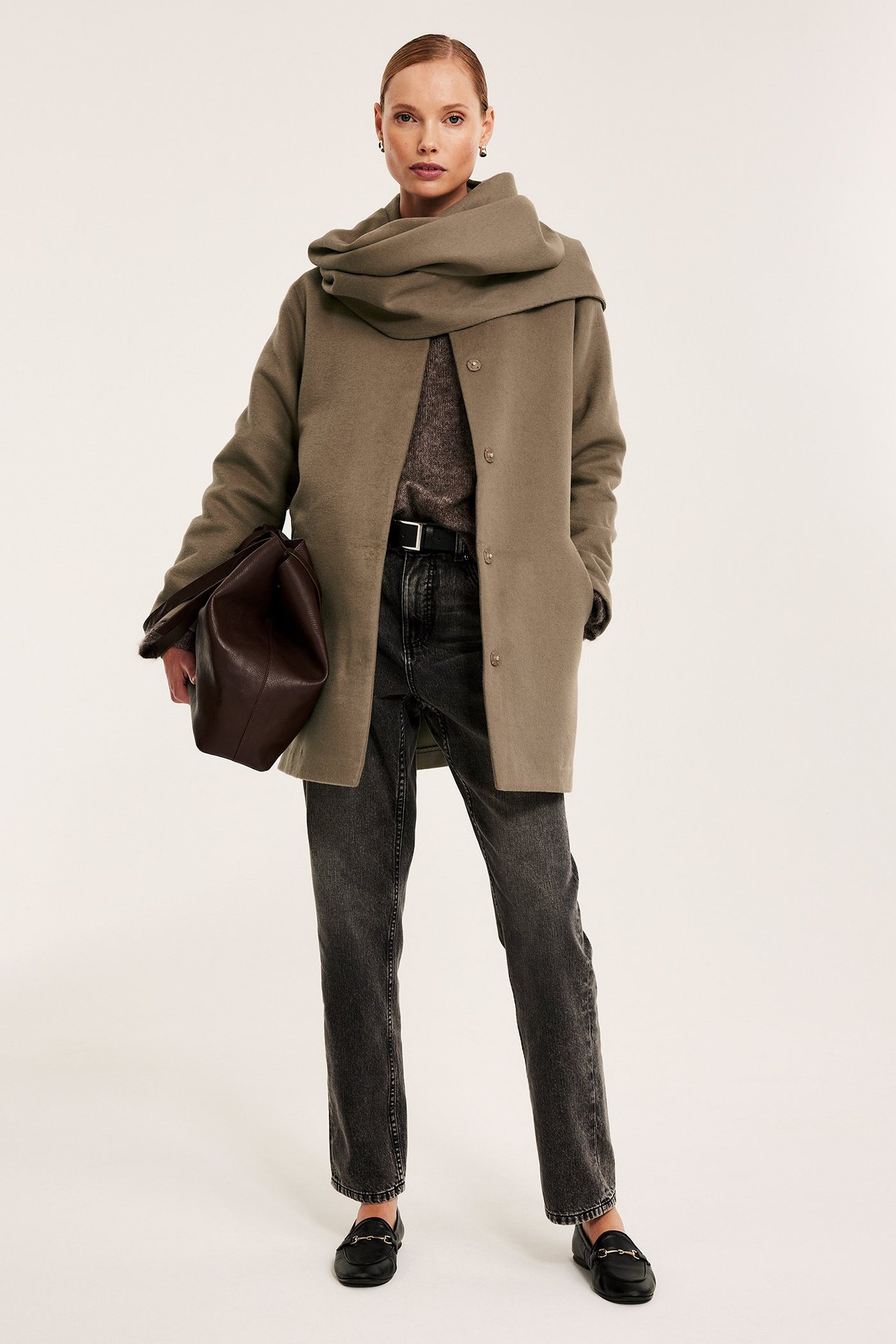 Edestä: nainen vaaleanruskeassa takissa huivikauluksella, tummissa farkuissa, mustissa loafer-kengissä, ruskea laukku kädessä.