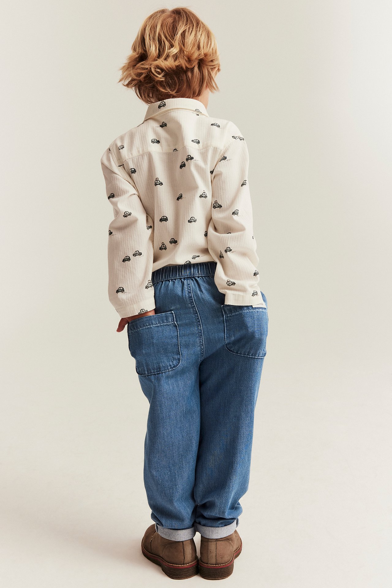 Ryggvy av ett barn i blå denimbyxor med resår i midjan och en ljus bilmönstrad skjorta.