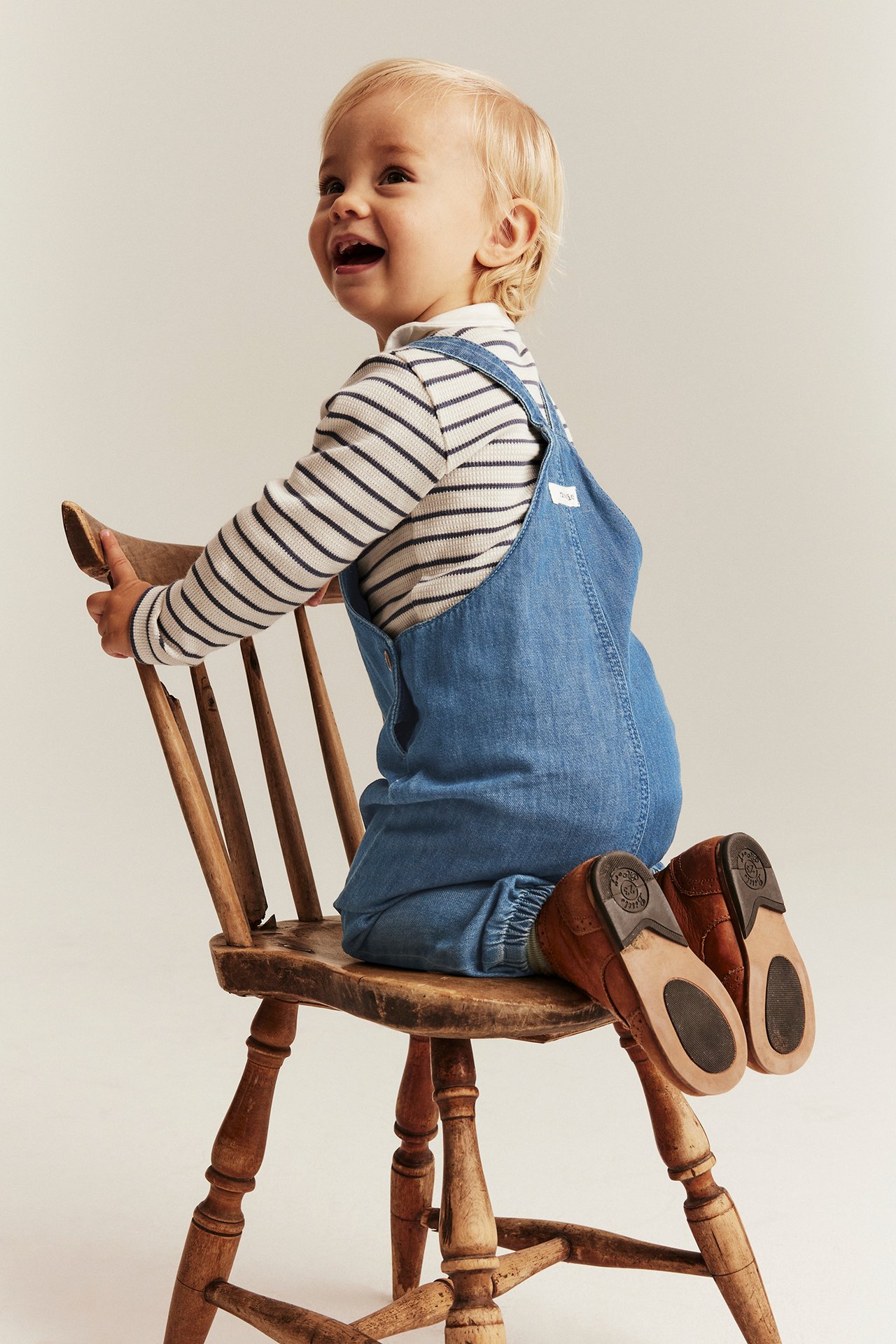 Z boku: dziecko w jasnoniebieskich dżinsowych ogrodniczkach z gumkami, pasiastym topie i brązowych butach.
