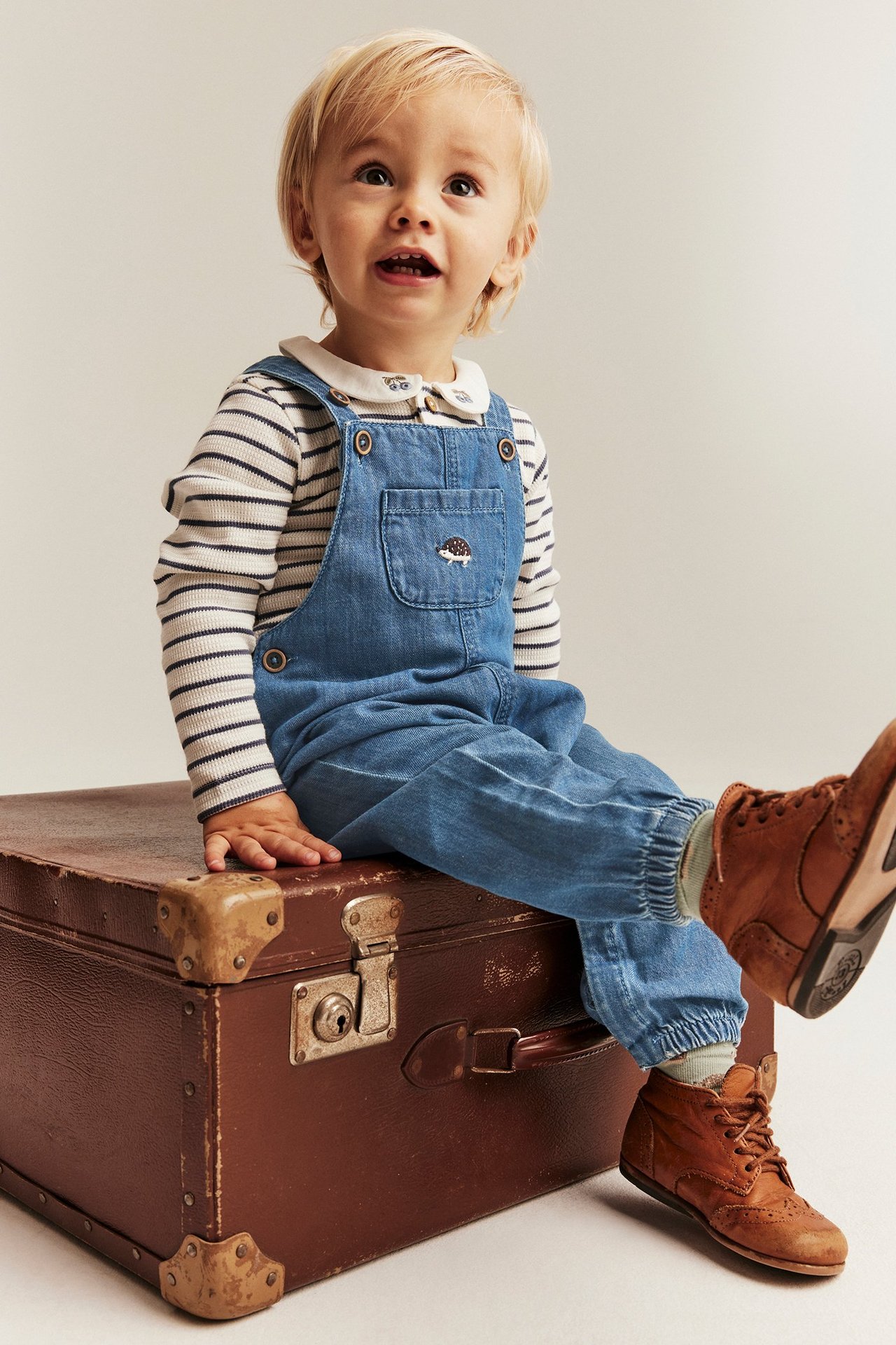 Vauva, edestä, siniset farkkuhaalarit siilikirjailulla, raidallinen paita ja ruskeat kengät jalassa.