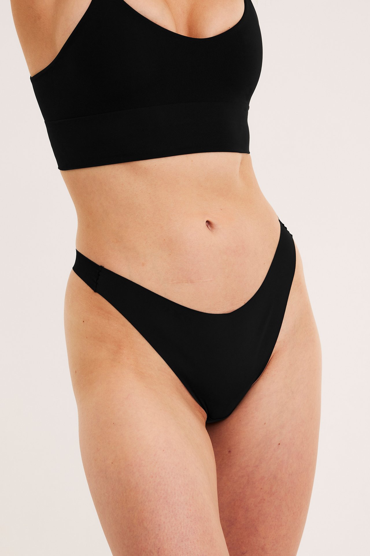 Frontutsikt av kvinne i svarte clean cut mikro stringtruser og en matchende svart bralette.