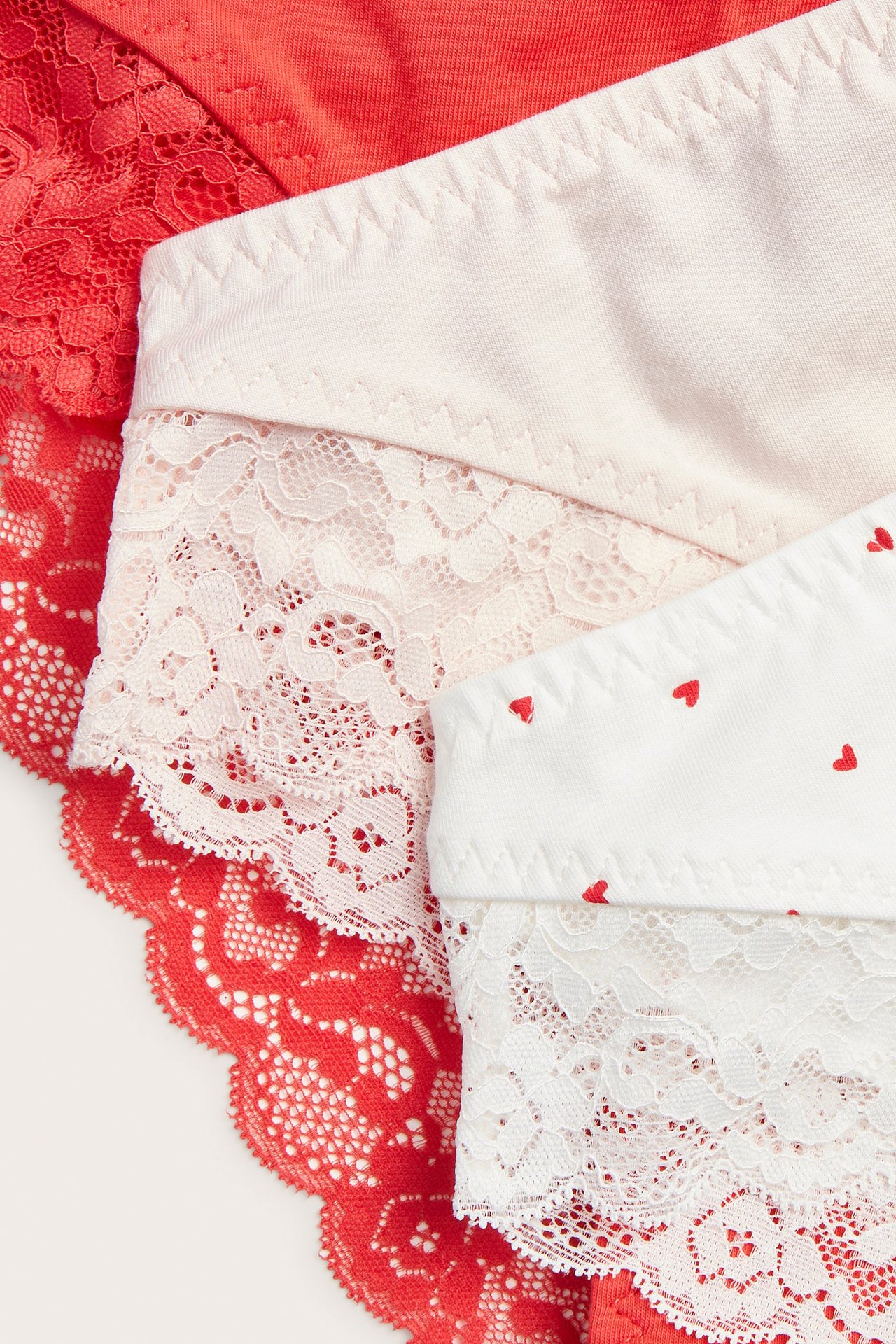 Nærbilde av tre midjehøye brazilian-truser: rød blonde, lys rosa blonde og hvit med rødt hjerteprint.