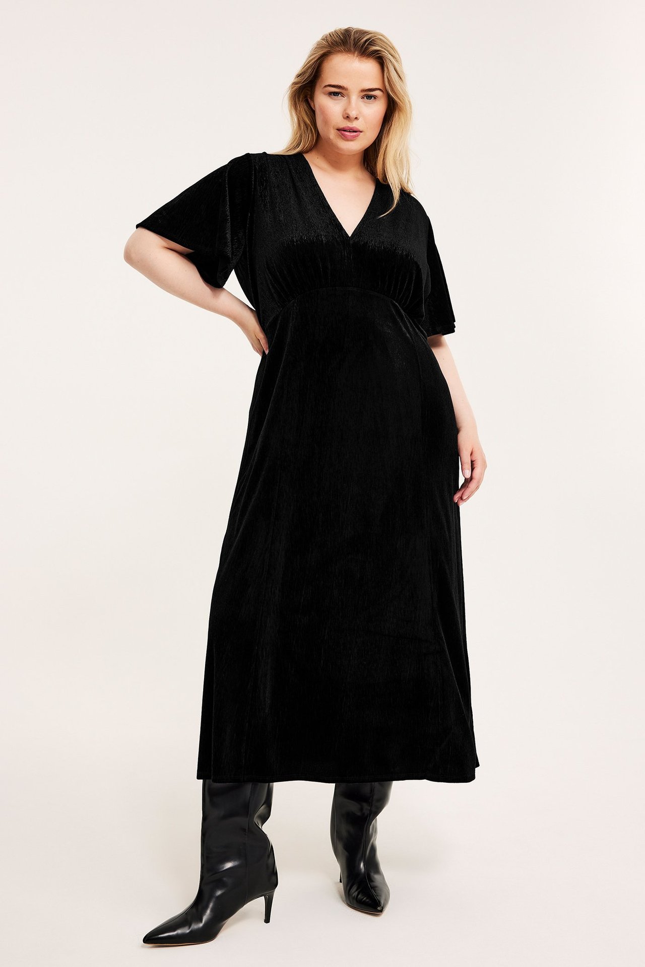 Edestäpäin nainen mustassa samettimekossa perhoshihoilla ja mustissa korkeakorkoisissa saappaissa.