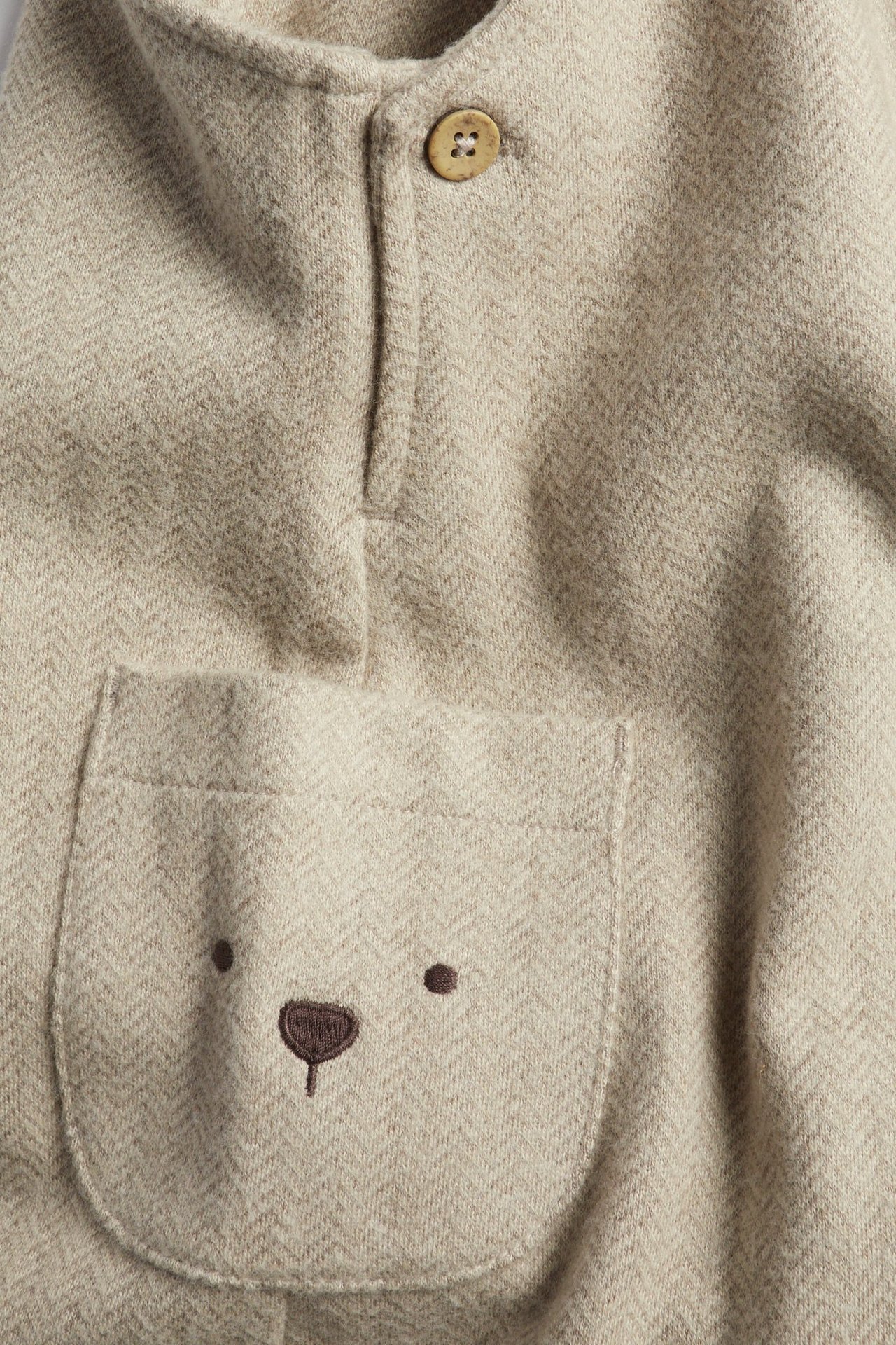 Lähikuva beige melange kalanruotokuosisista haalareista, joissa puunappi ja tasku brodeeratulla karhun kasvoilla.