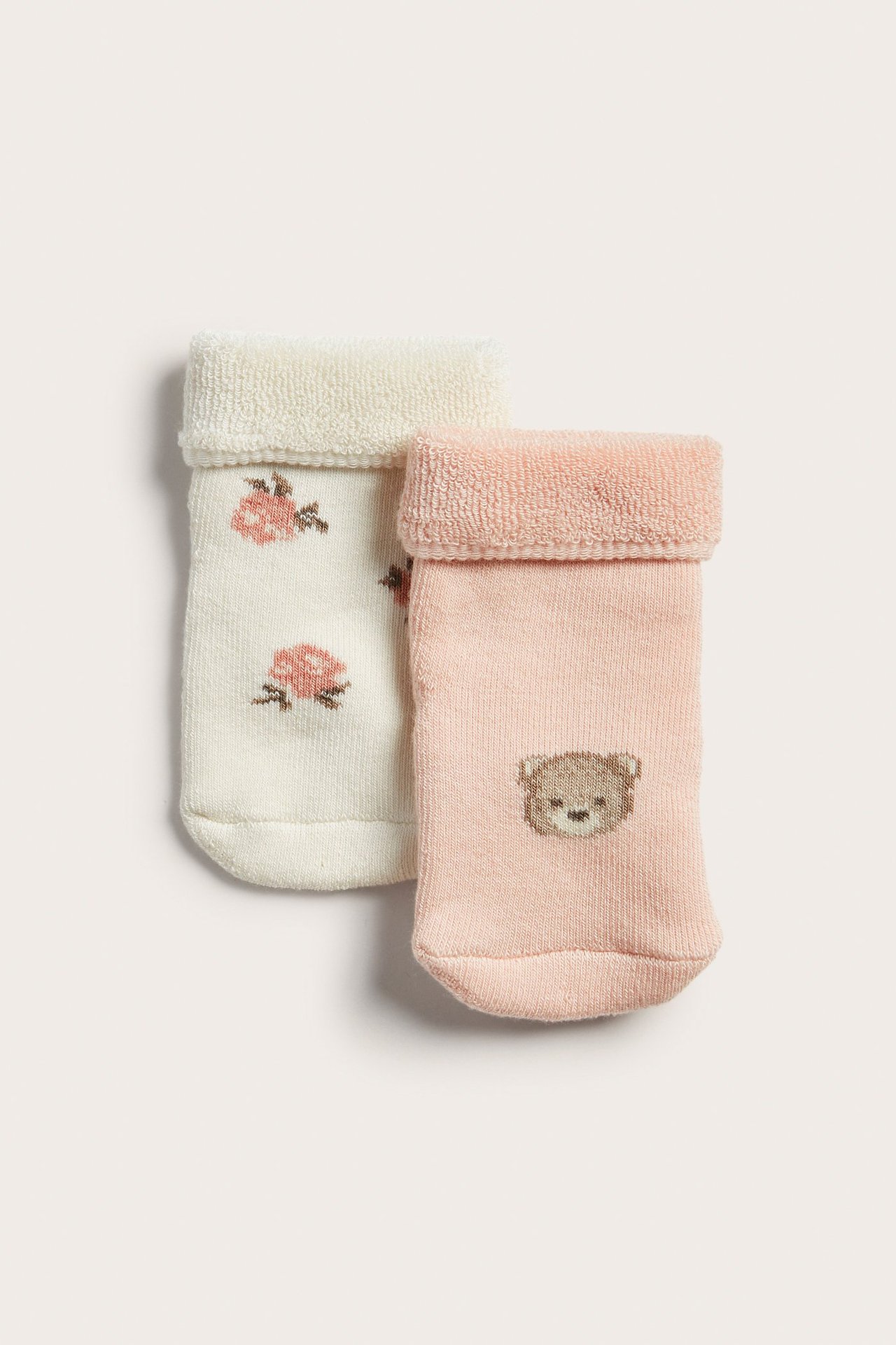 Nærbilde forfra av to par babyfrottésokker: lys rosa med teddybjørn, kremhvite med roser.