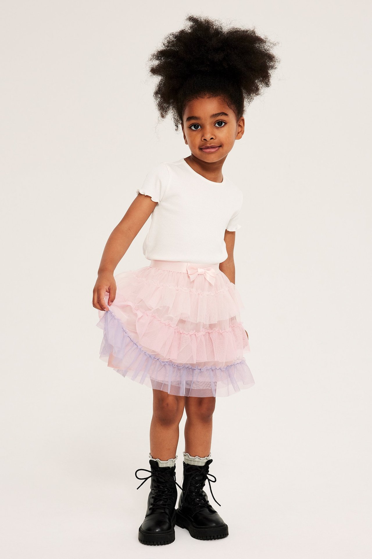 Frontvy: Barn i ljusrosa och lila volangkjol i tyll med rosett, vit ribbad topp och svarta kängor.