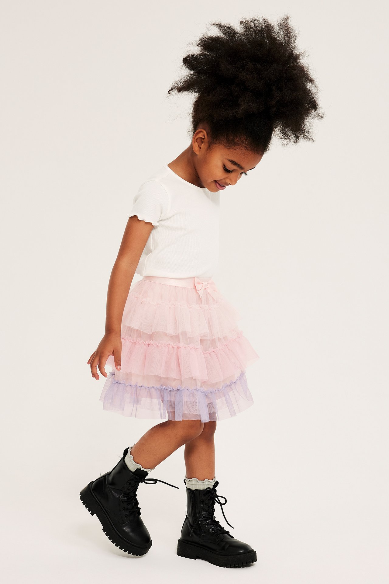 Sidovy: Barn i ljusrosa och lila volangkjol i tyll, vit t-shirt och svarta kängor.