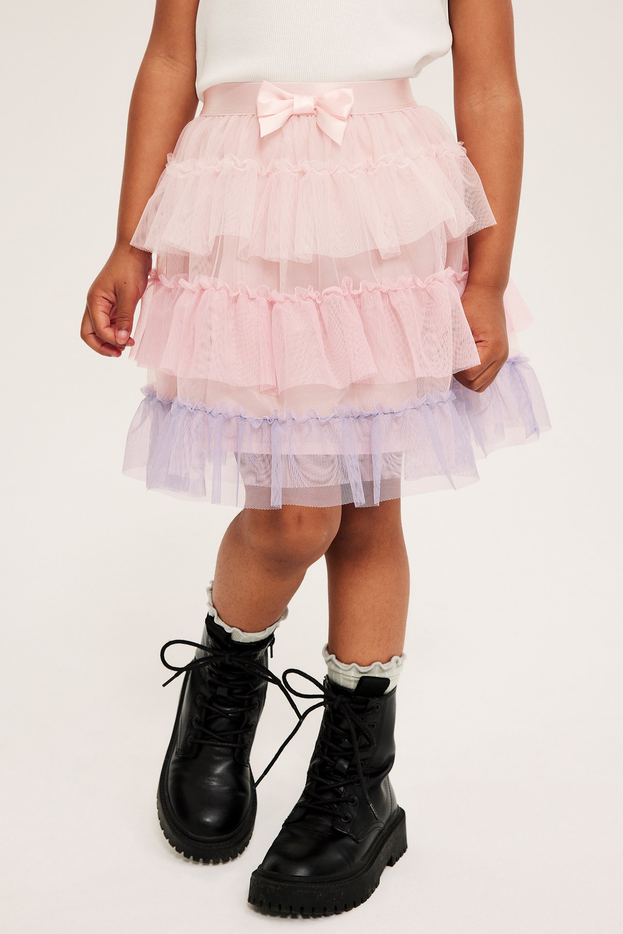 Framifrån: Barn bär ljusrosa tyllkjol med rosett, vit topp, svarta kängor, vita strumpor.