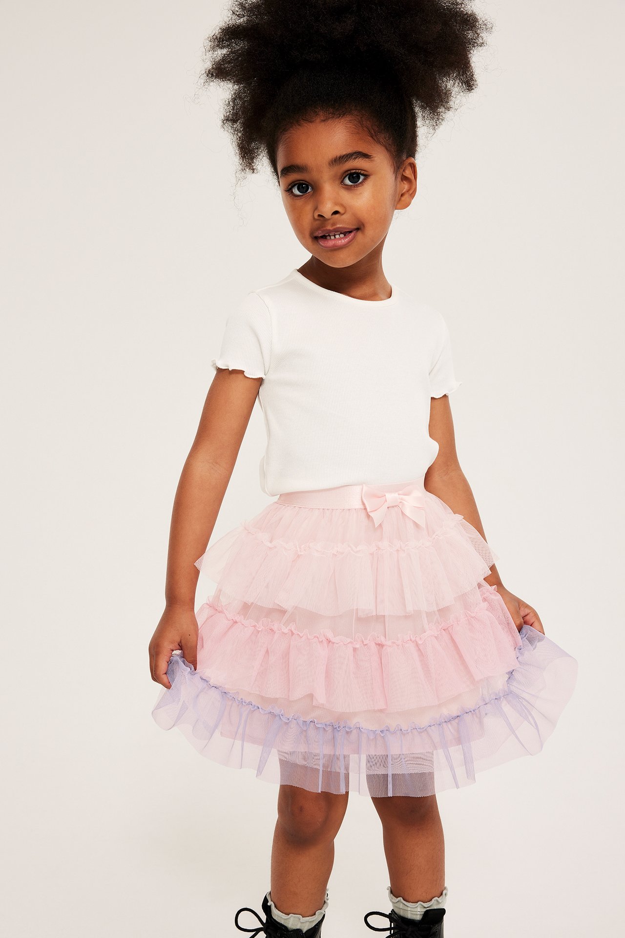 Framifrån: Kid i ljusrosa tyllkjol med volanger och lila kant, samt en vit ribbad topp.