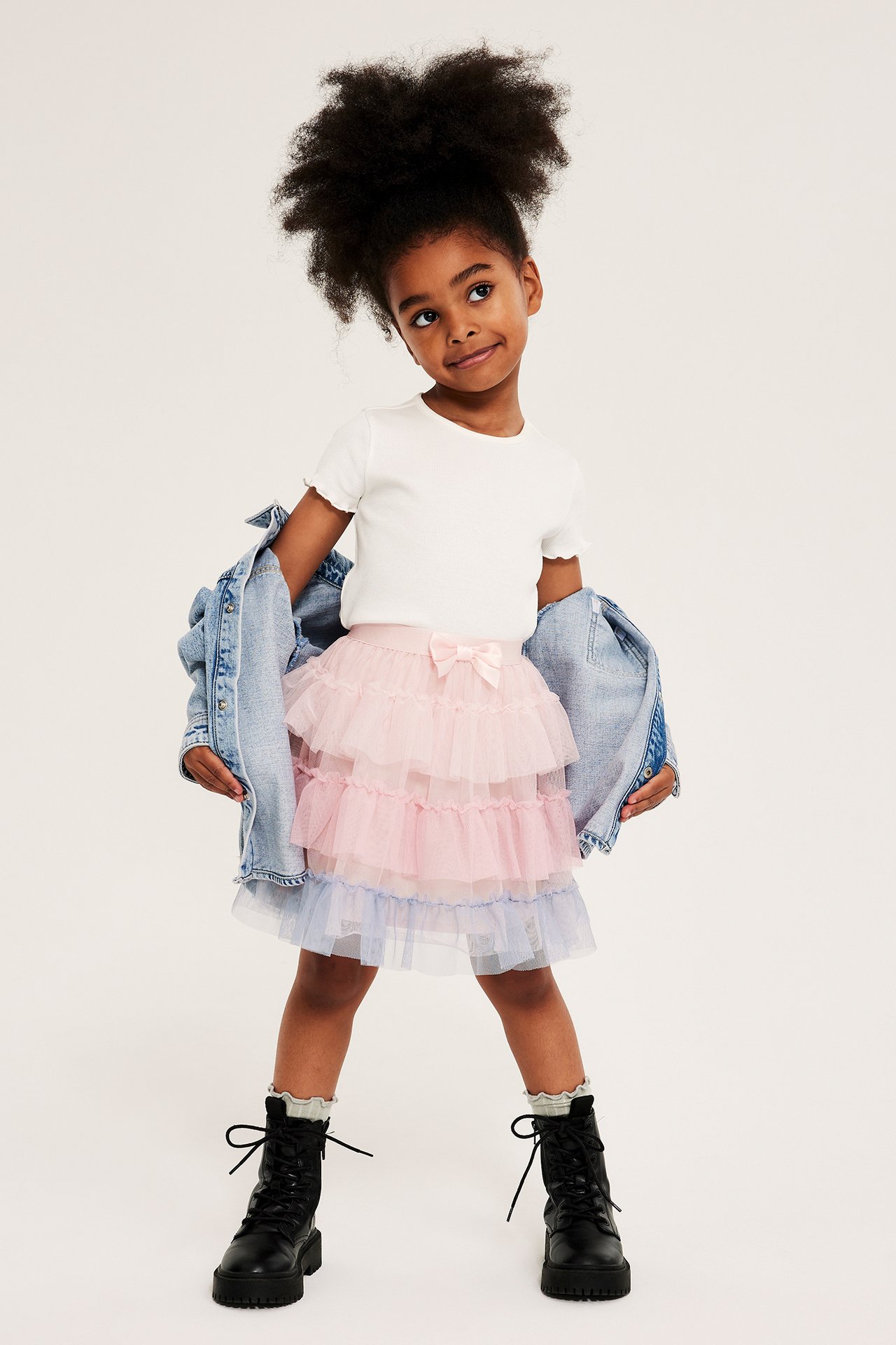 Widok z przodu: Dziecko ma jasnoróżowo-niebieską tiulową spódniczkę z kokardą, biały t-shirt, kurtkę jeansową, czarne buty.