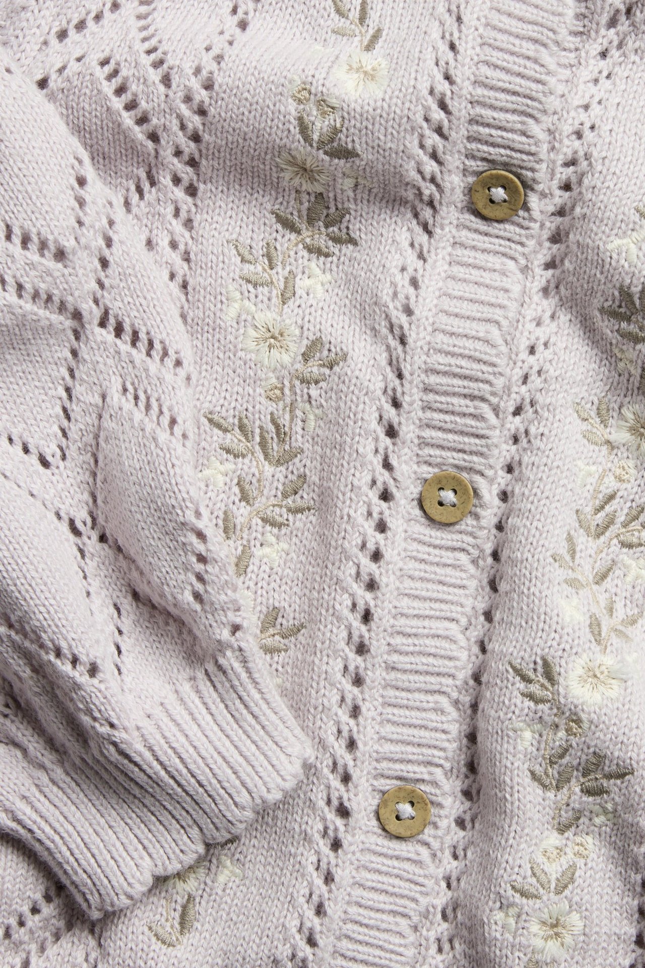 Närbild på en ljuslila mönsterstickad bomullskofta med broderade blommor och träknappar.