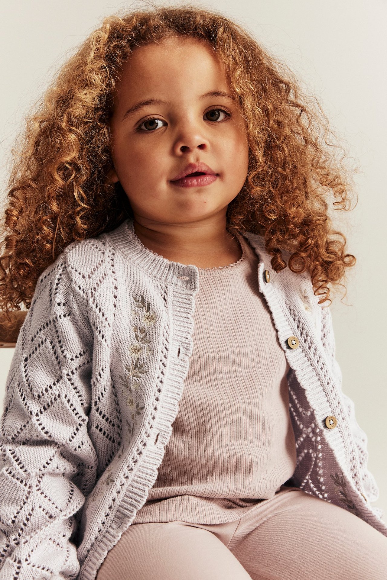 Edestä: Lapsi vaaleanlilaa kuvioneulottua puuvillatakkia kukkakirjonnalla, ribbipaidan ja leggingsien päällä.