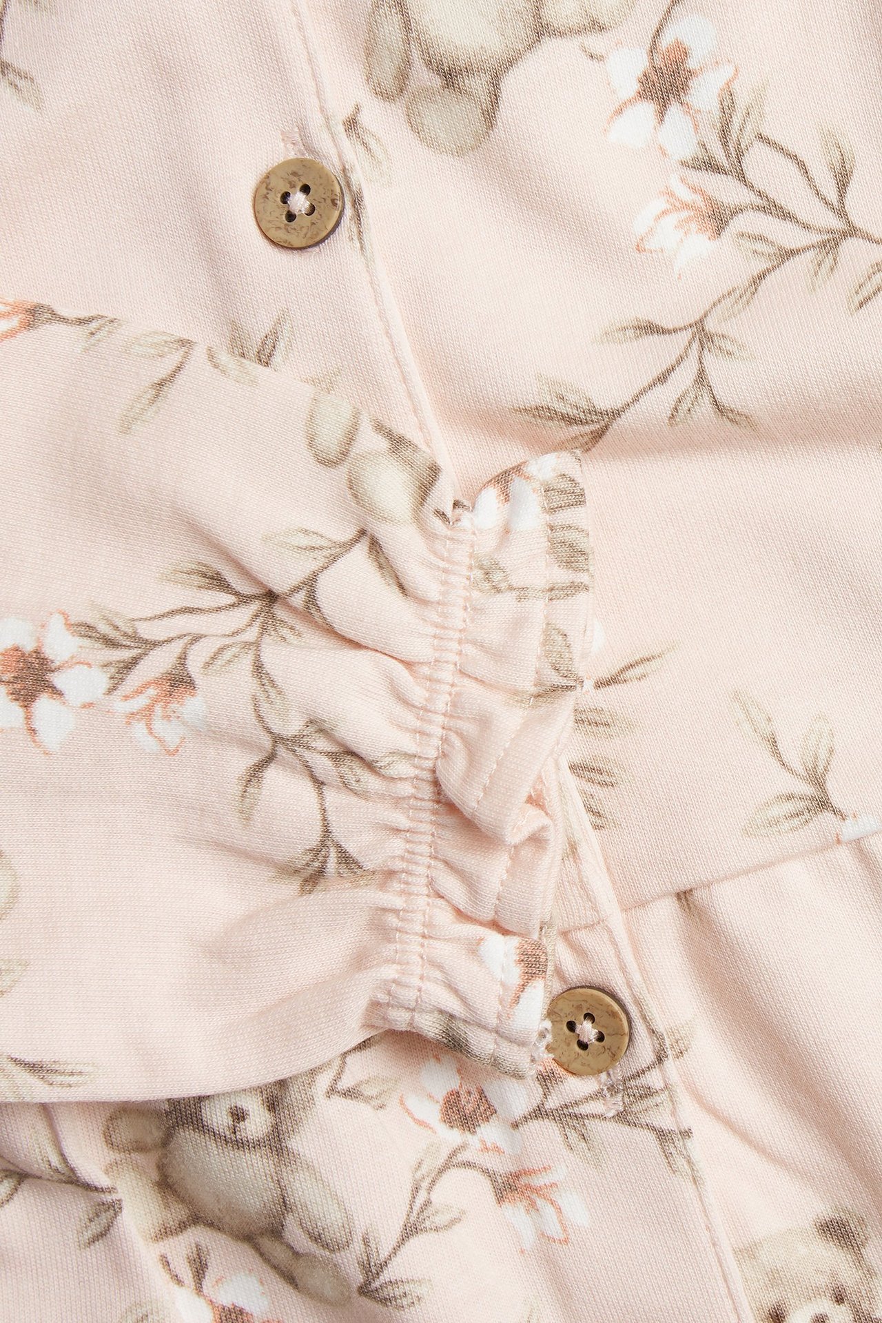 Nærbilde av en lys rosa baby jumpsuit i bomull med blomster- og bjørnemønster, volanger og treknapper.