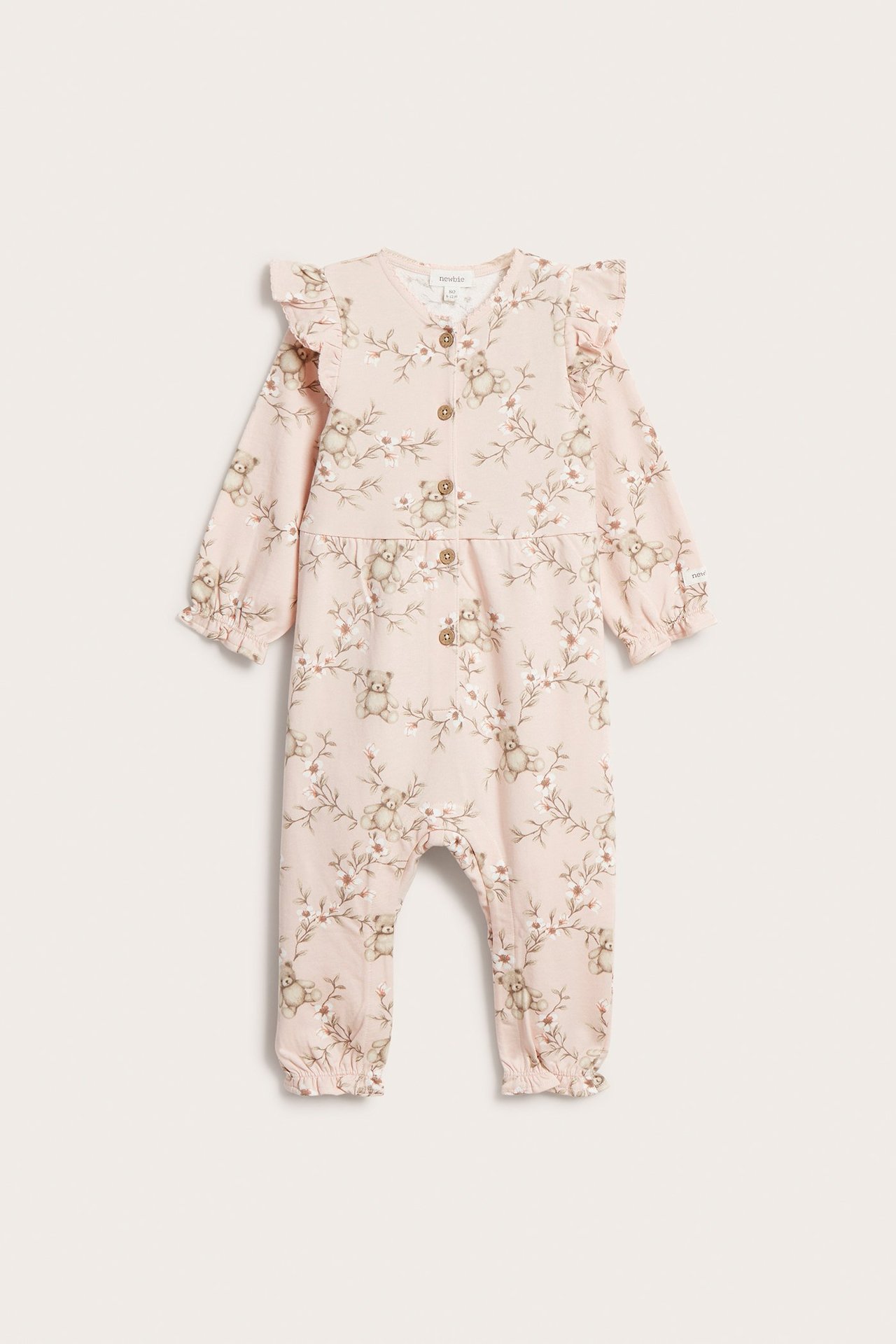 Frontvisning av en lyserosa babyjumpsuit i bomull med blomster- og bamsemønster, volanger og knapper.