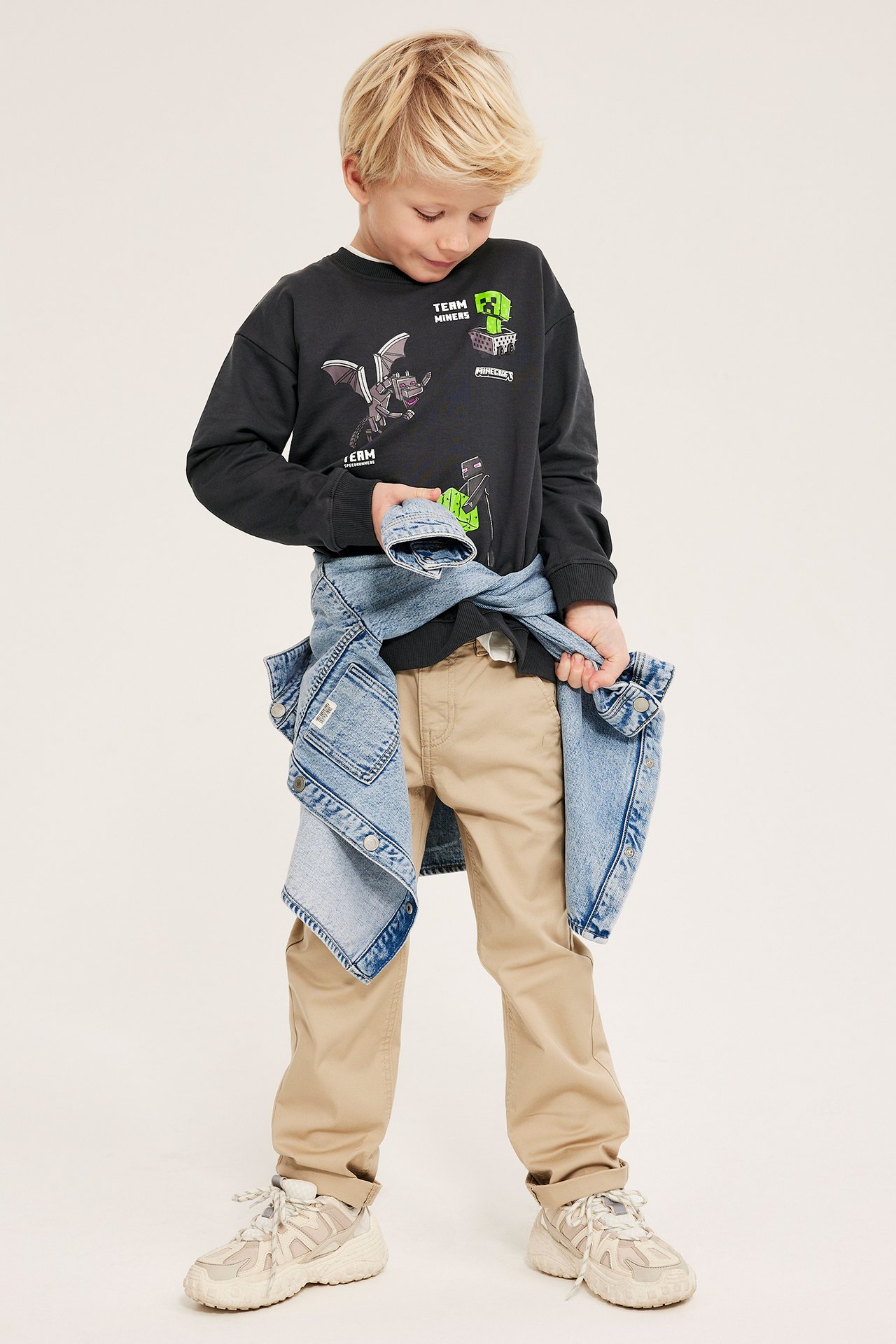 Edestäpäin: lapsi mustassa Minecraft-paidassa, beigenvärisissä housuissa, vaalea farkkutakki vyötäröllä.