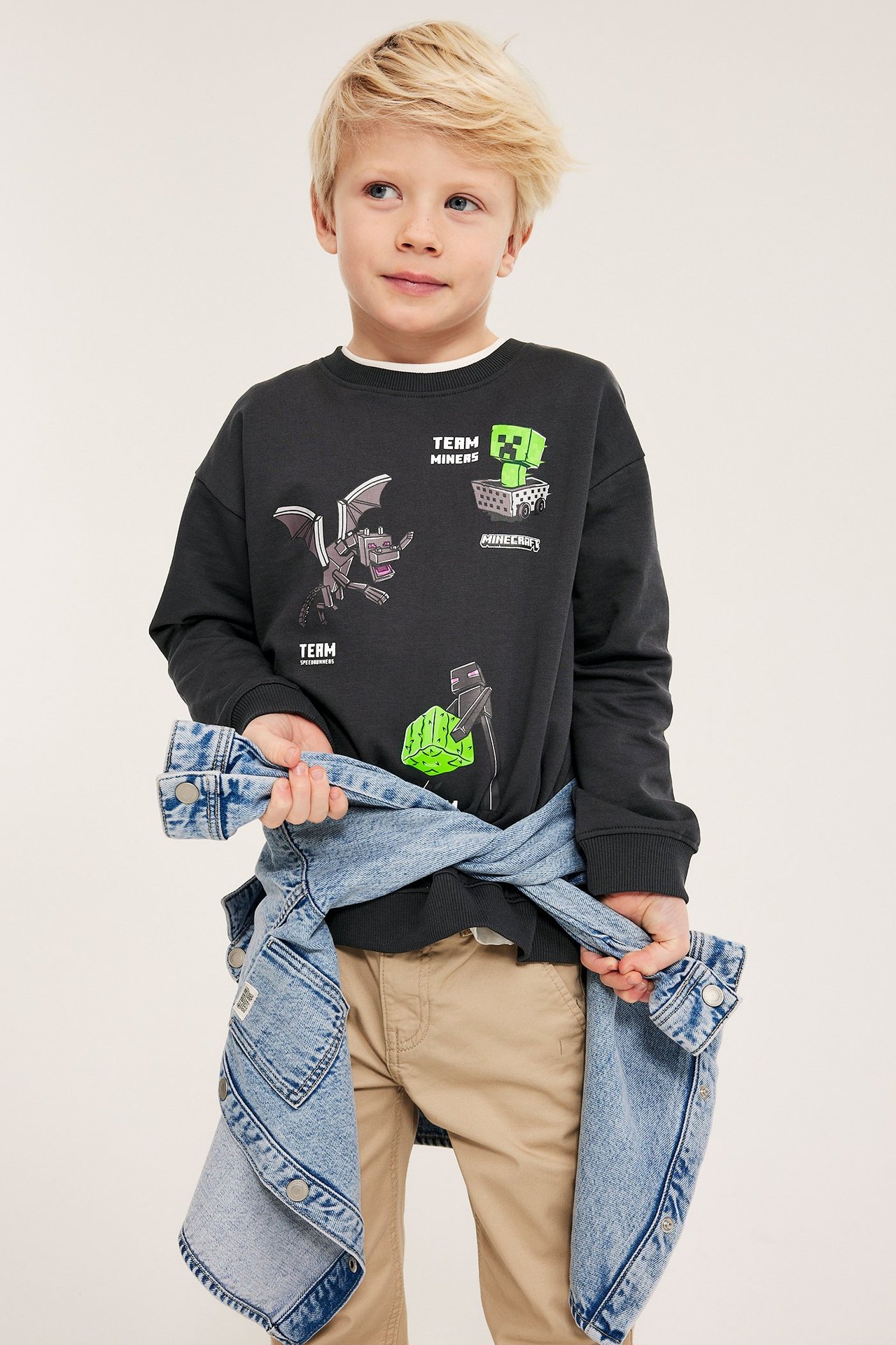 Edestäpäin, lapsi yllään musta puuvillainen Minecraft-painatuksella varustettu collegepaita, beiget housut ja vyötäröllä farkkutakki.
