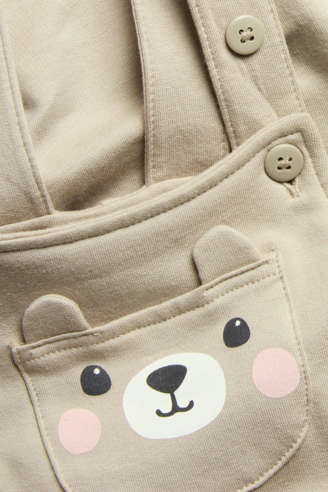 Zbliżenie beżowych ogrodniczek niemowlęcych z nadrukiem twarzy misia i uszami na kieszeni, z zapinanymi szelkami.