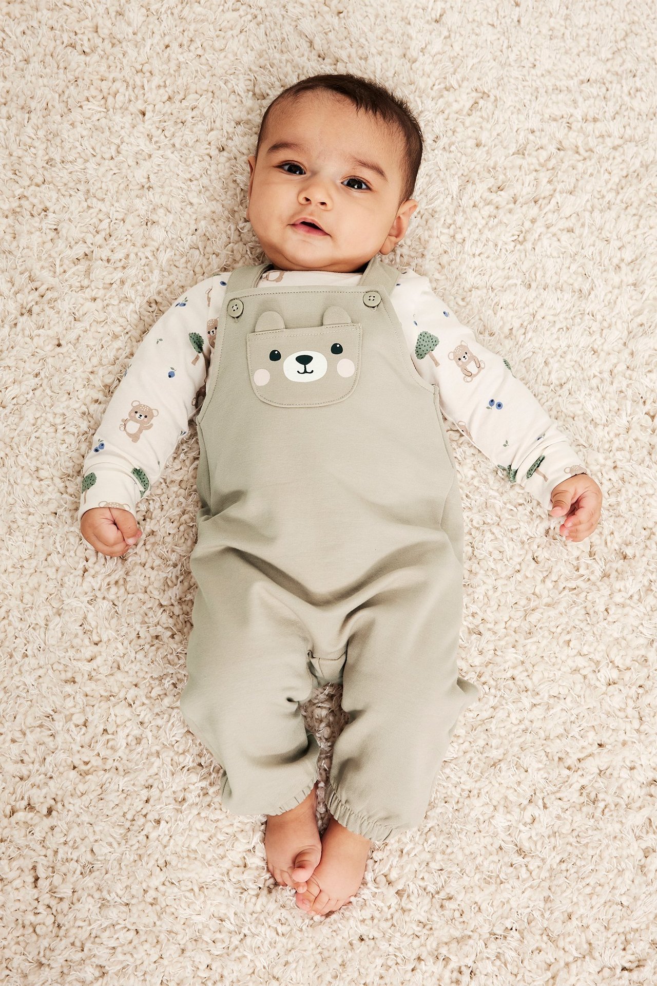 Widok z góry na dziecko w beżowych ogrodniczkach z kieszenią w kształcie misia na białej bluzce w wzory.