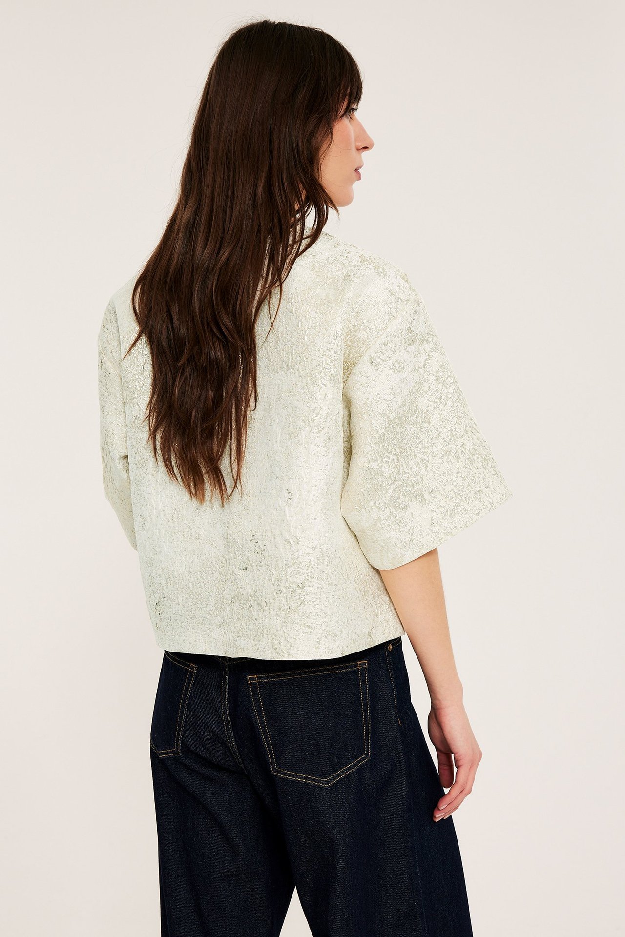 Bakfra-sidebilde av kvinne i offwhite jacquardbluse med korte ermer og mørkeblå jeans.
