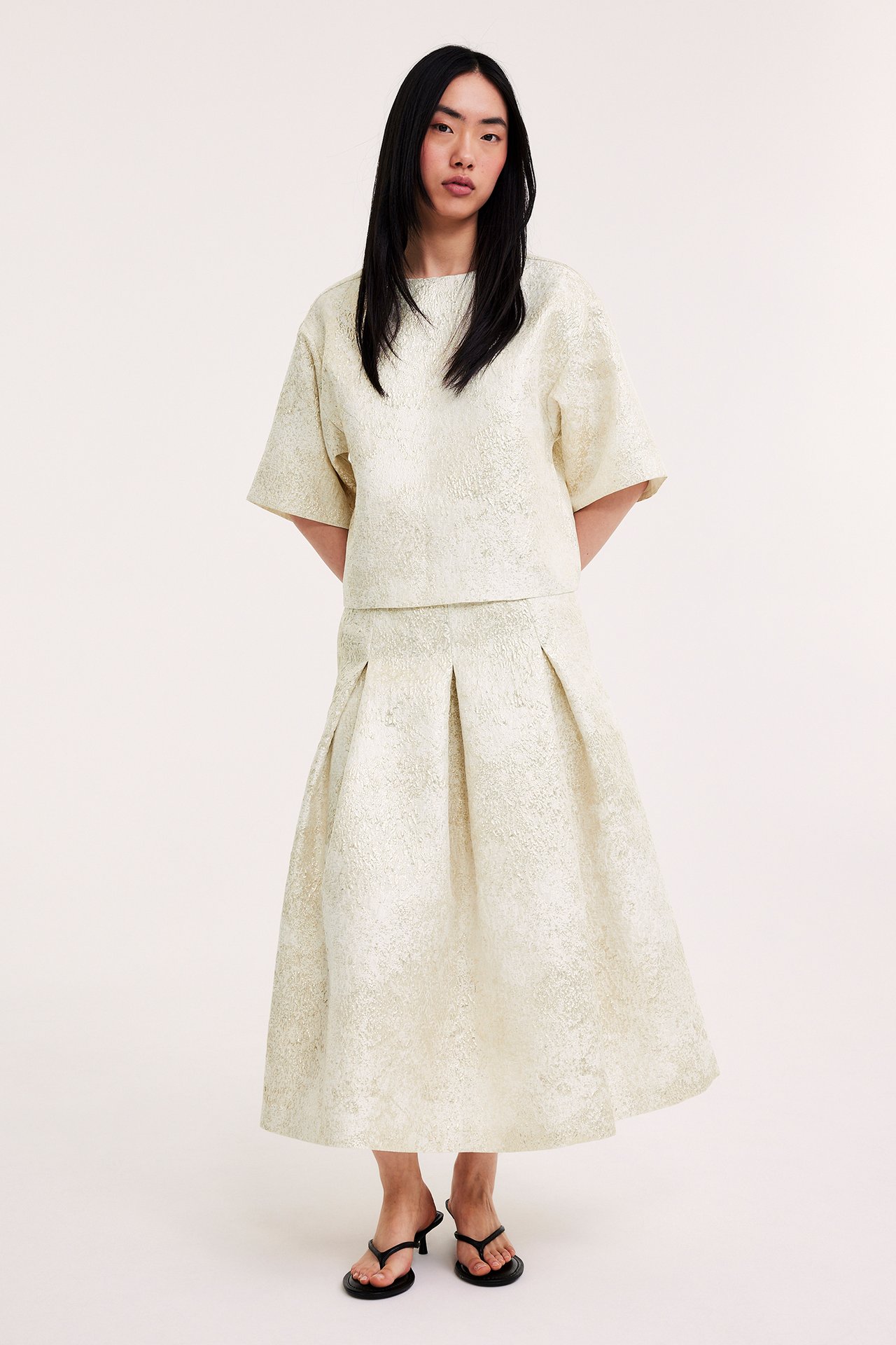 Edestäpäin nainen yllään luonnonvalkoinen jacquardpusero, yhteensopiva laskostettu hame ja mustat varvassandaalit.