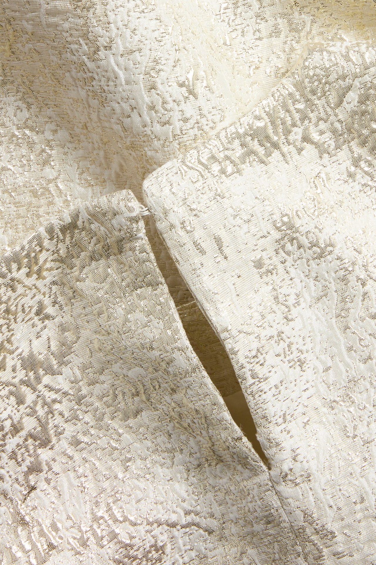Nærbilde av en offwhite brokadebluse med gulldetaljert jacquardmønster og knappelukking.