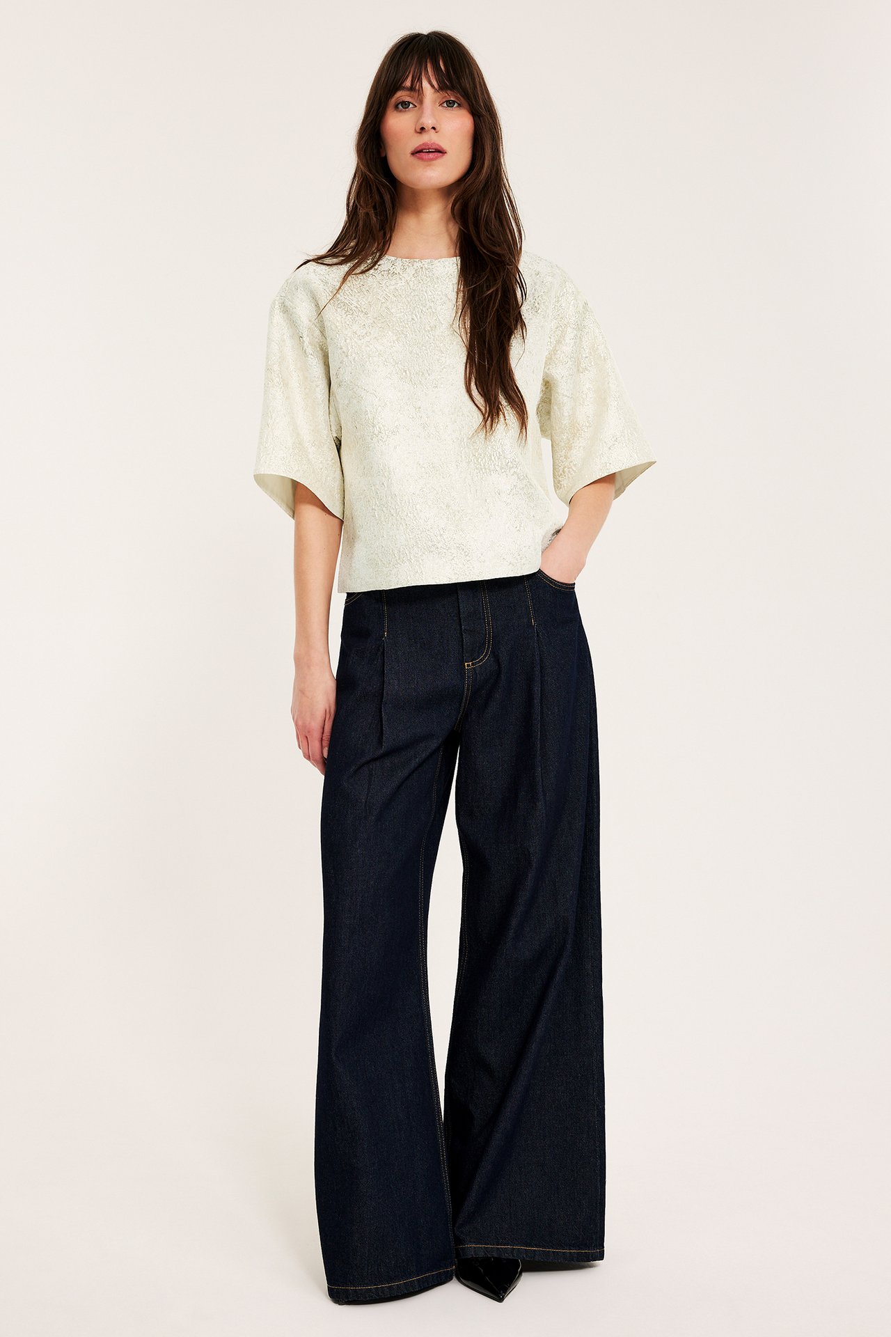 Frontutsikt av kvinne i en offwhite jacquardmønstret bluse med vide ermer og mørkeblå vide jeans.