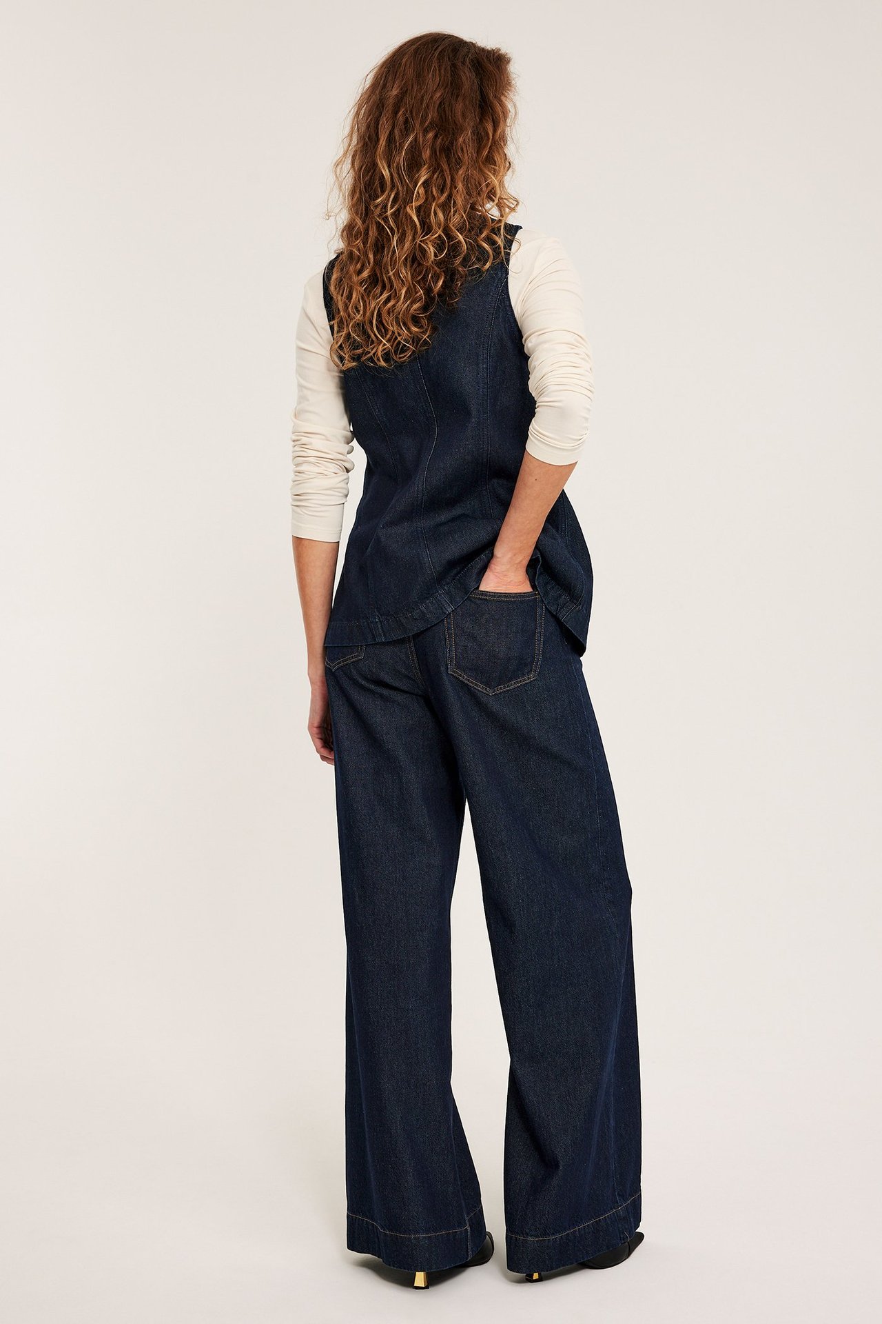 Kvinna bakifrån i mörkblå vida jeans med sömmar framtill, denimväst och ljusbeige topp.