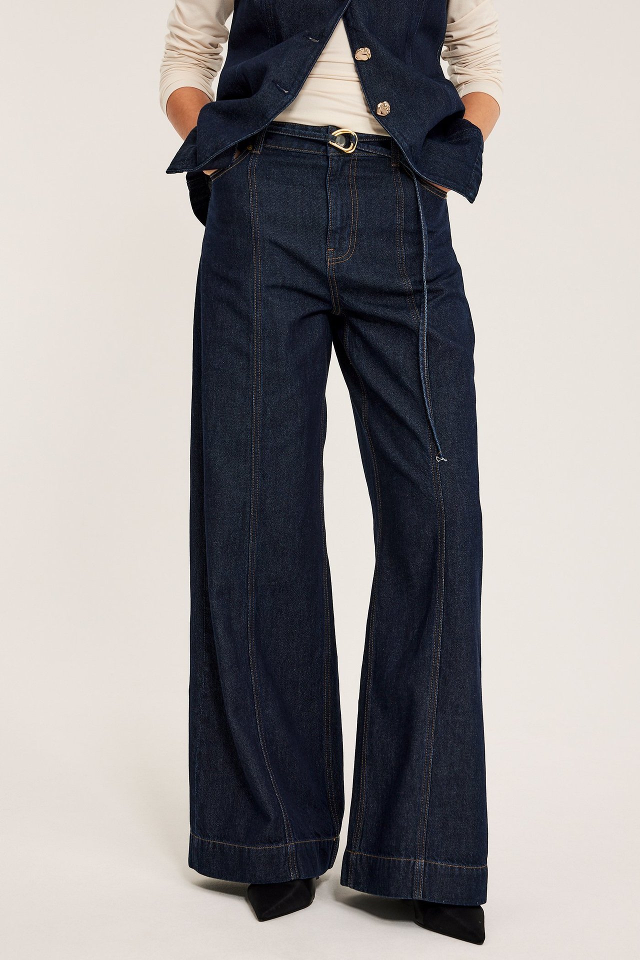 Etunäkymä naisesta yllään tummansiniset leveälahkeiset korkeavyötäröiset farkut etusaumoilla ja kultasolkinen vyö.