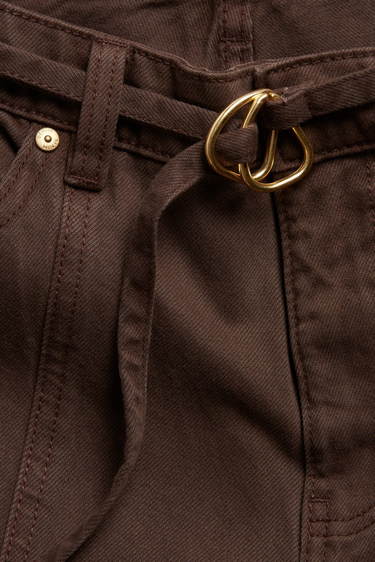 Nærbilde forfra av mørkebrune vide high-waist jeans med matchende stoffbelte og gullfarget dobbelspenne.