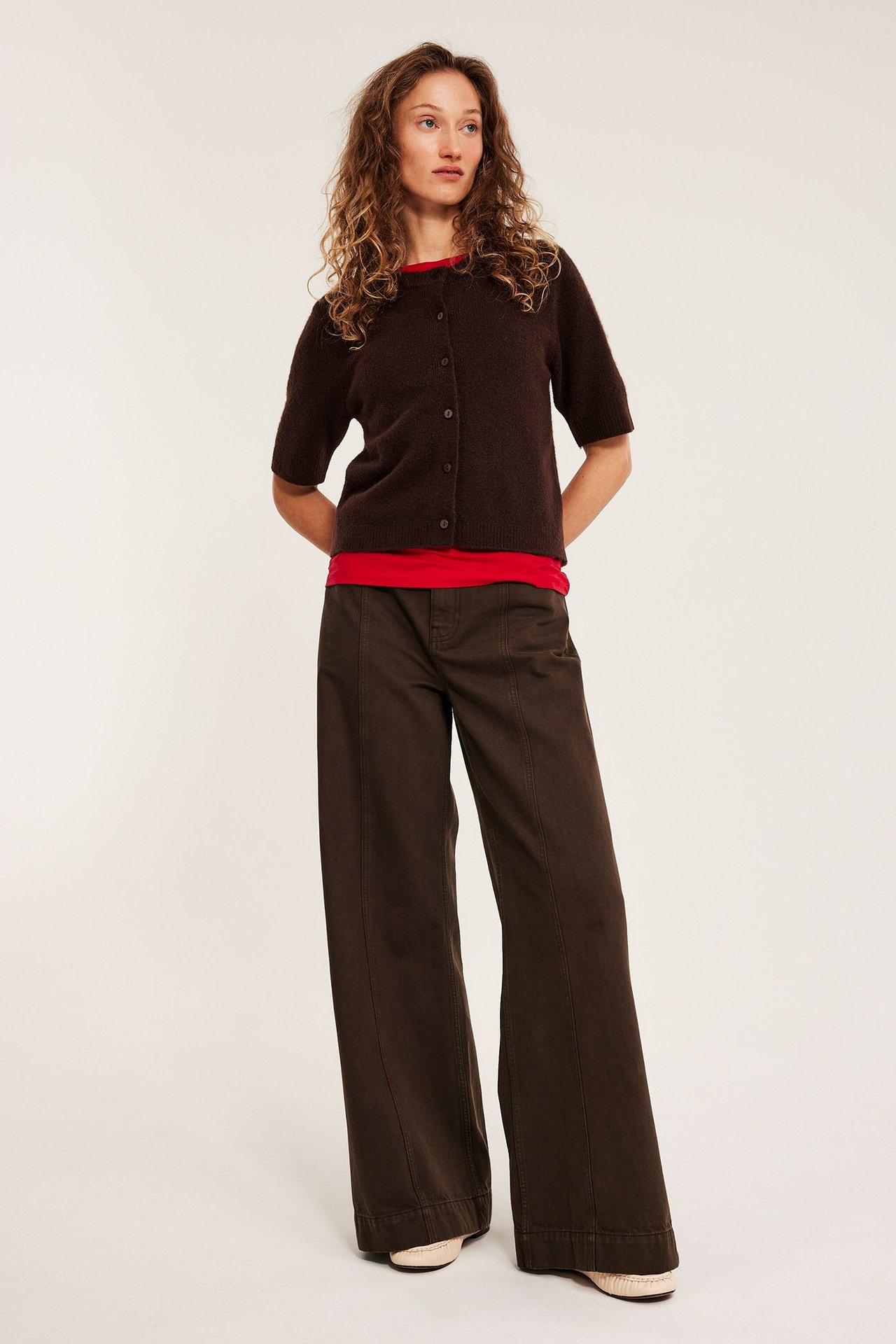Frontbilde av kvinne i mørkebrune vide jeans med sømmer foran, rød t-skjorte og brun cardigan.