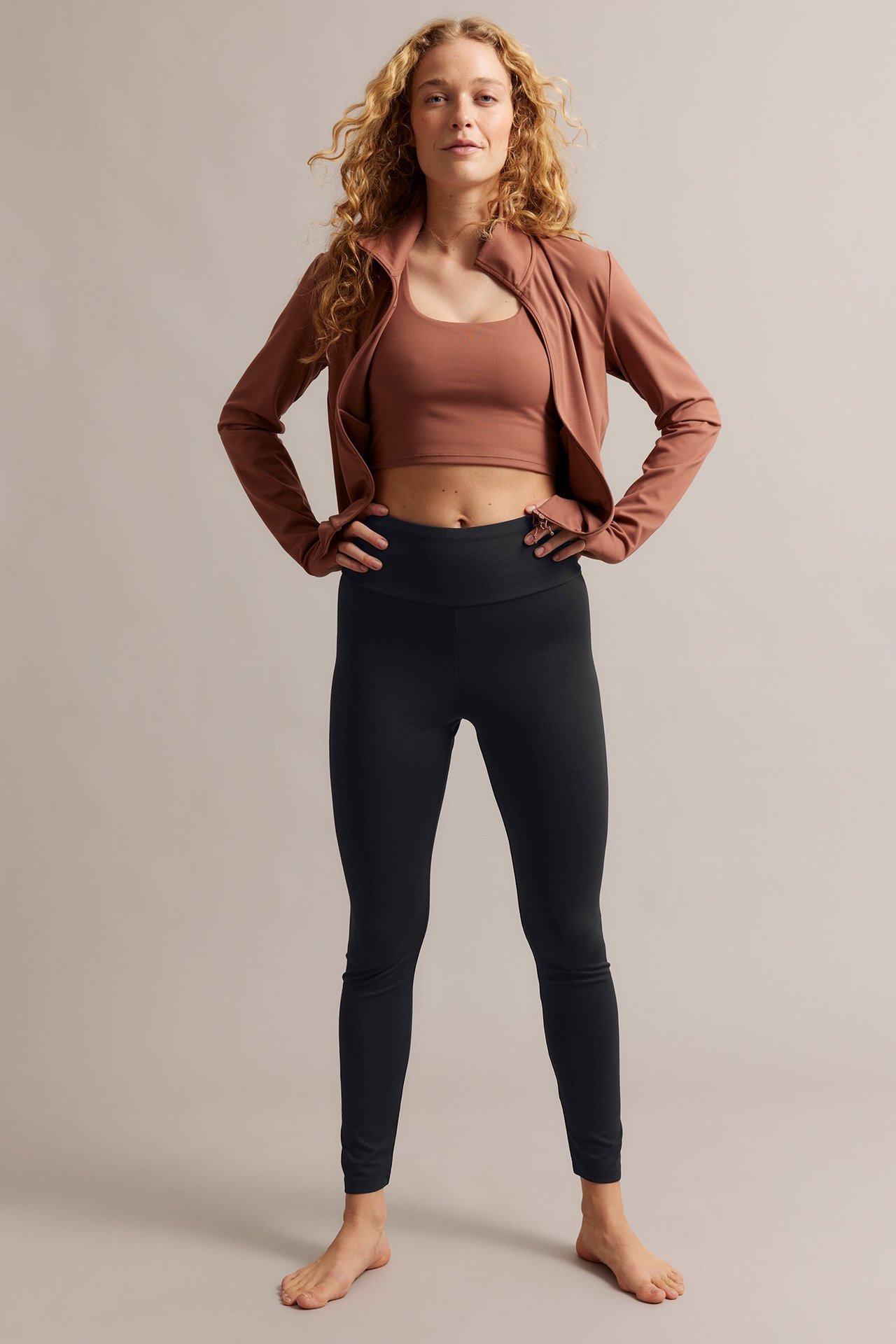 Frontbilde: Kvinne i svarte leggings med høyt liv, en rustfarget crop top og en åpen rustfarget jakke.
