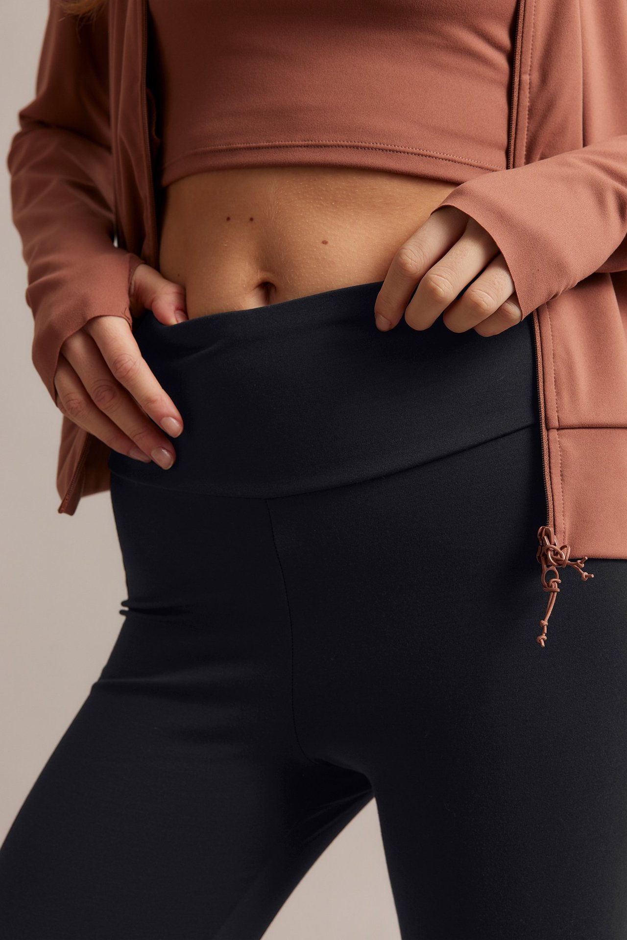 Närbild framifrån på kvinna i svarta leggings med hög midja, brunröd kort topp och öppen jacka.