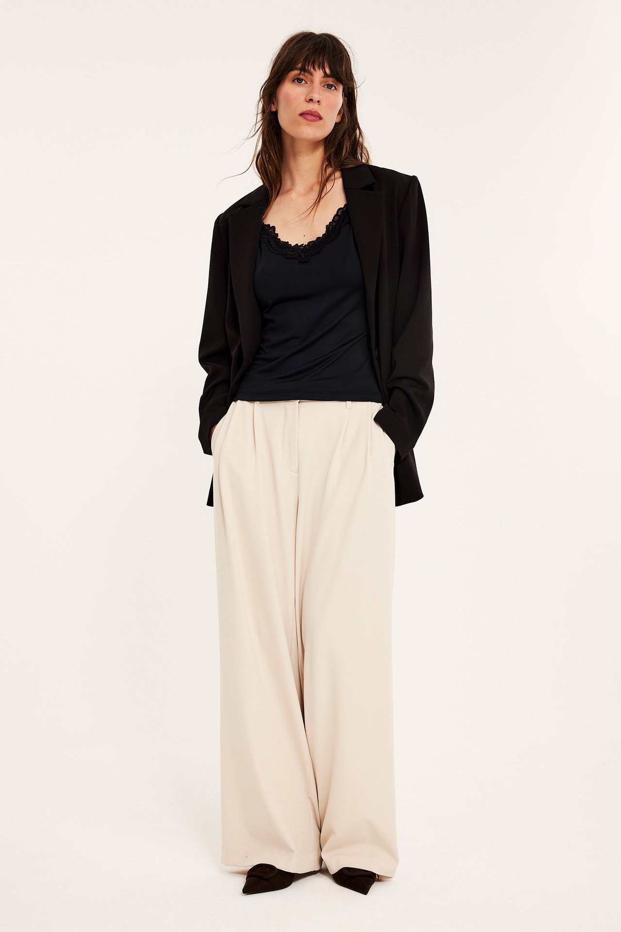 Forfra: kvinne i svart blonde-topp, svart blazer og lysebeige vide bukser.