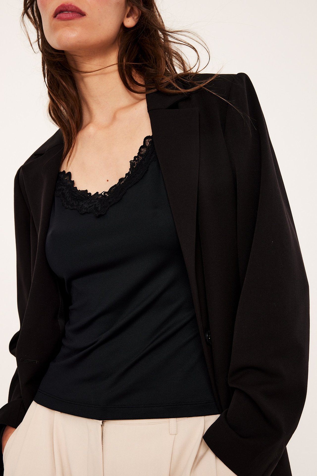 Forfra: Kvinne i svart blondetopp, åpen svart blazer og lysebeige bukser.