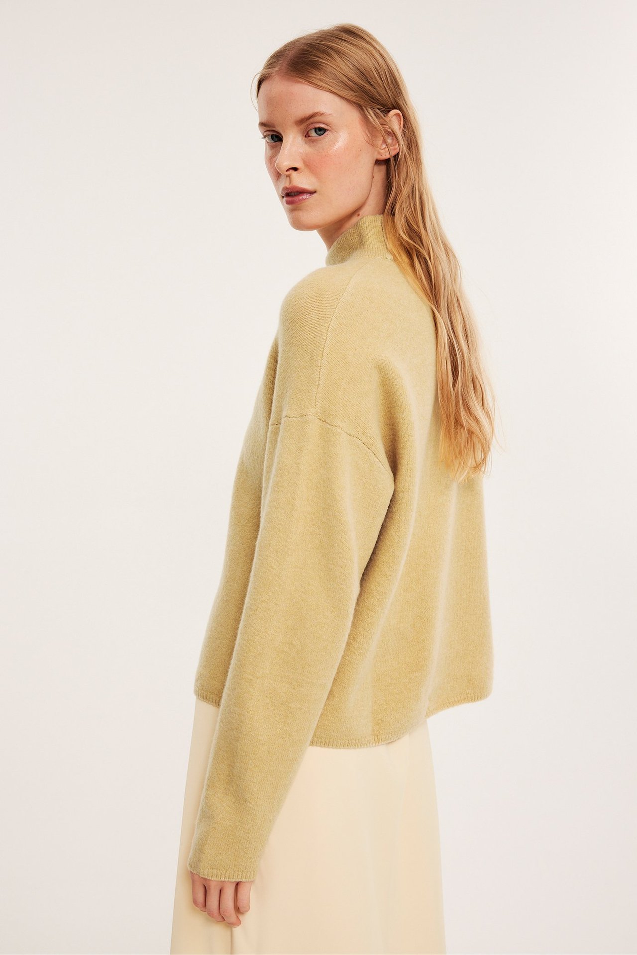 Sidebilde av en kvinne i en lys gul strukturert turtleneck-genser og et lys beige skjørt.